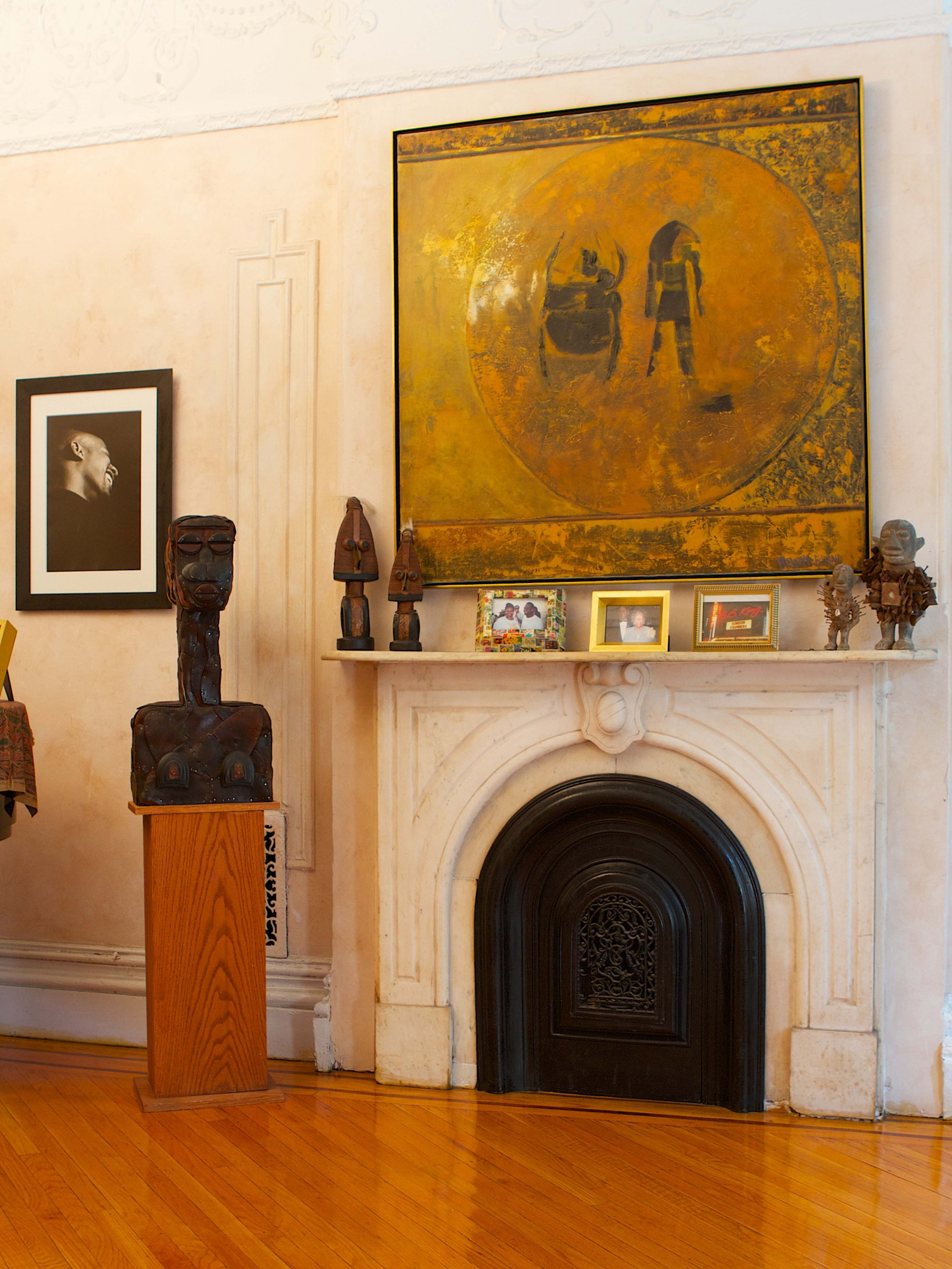 A fireplace with an ornate mantle adorned with a large painting, framed photographs, and wooden sculptures in a room with light-colored walls and polished wooden flooring.