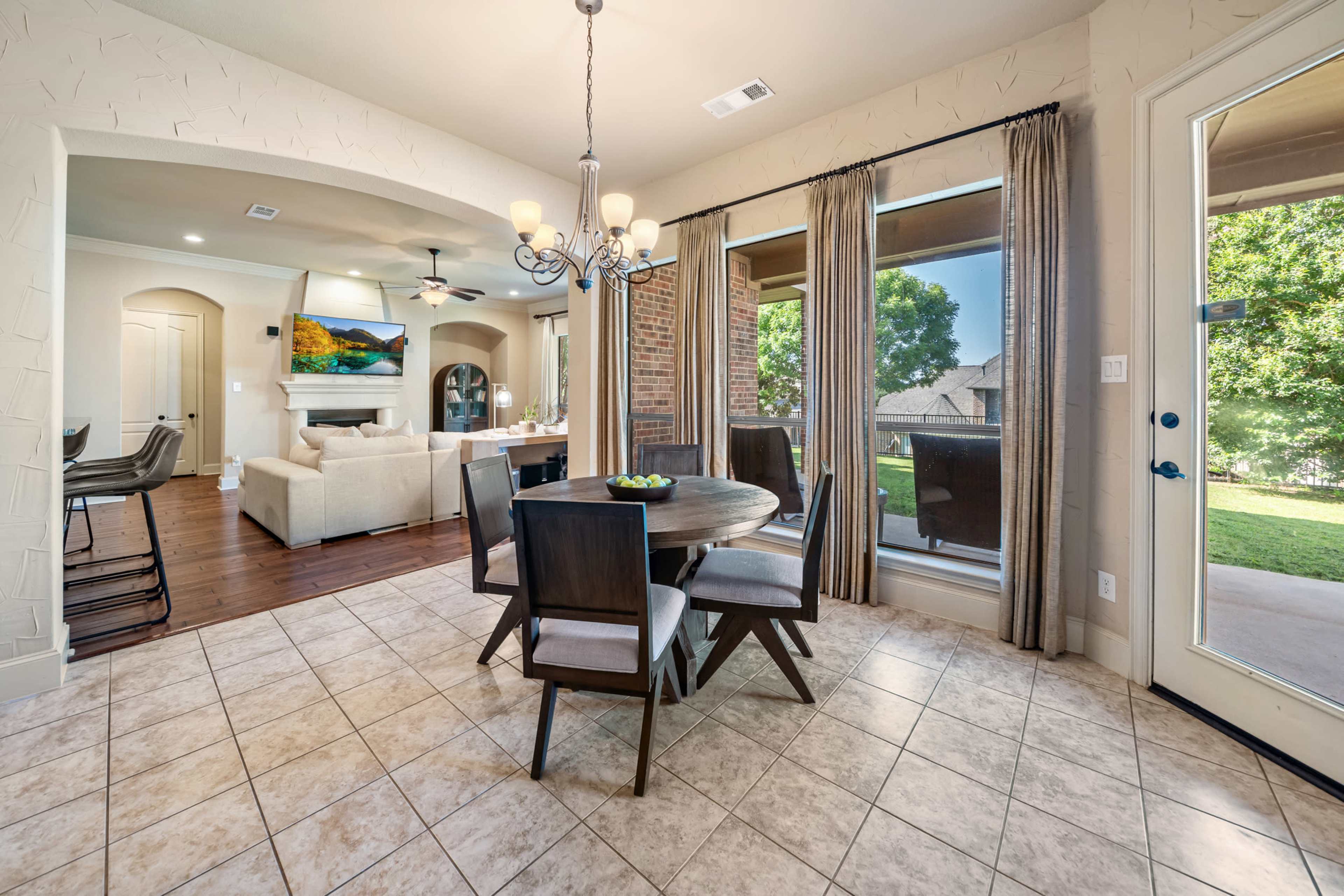 A dining area features a round table with six chairs, surrounded by large windows that overlook a grassy outdoor space, and is adjacent to an open living room.