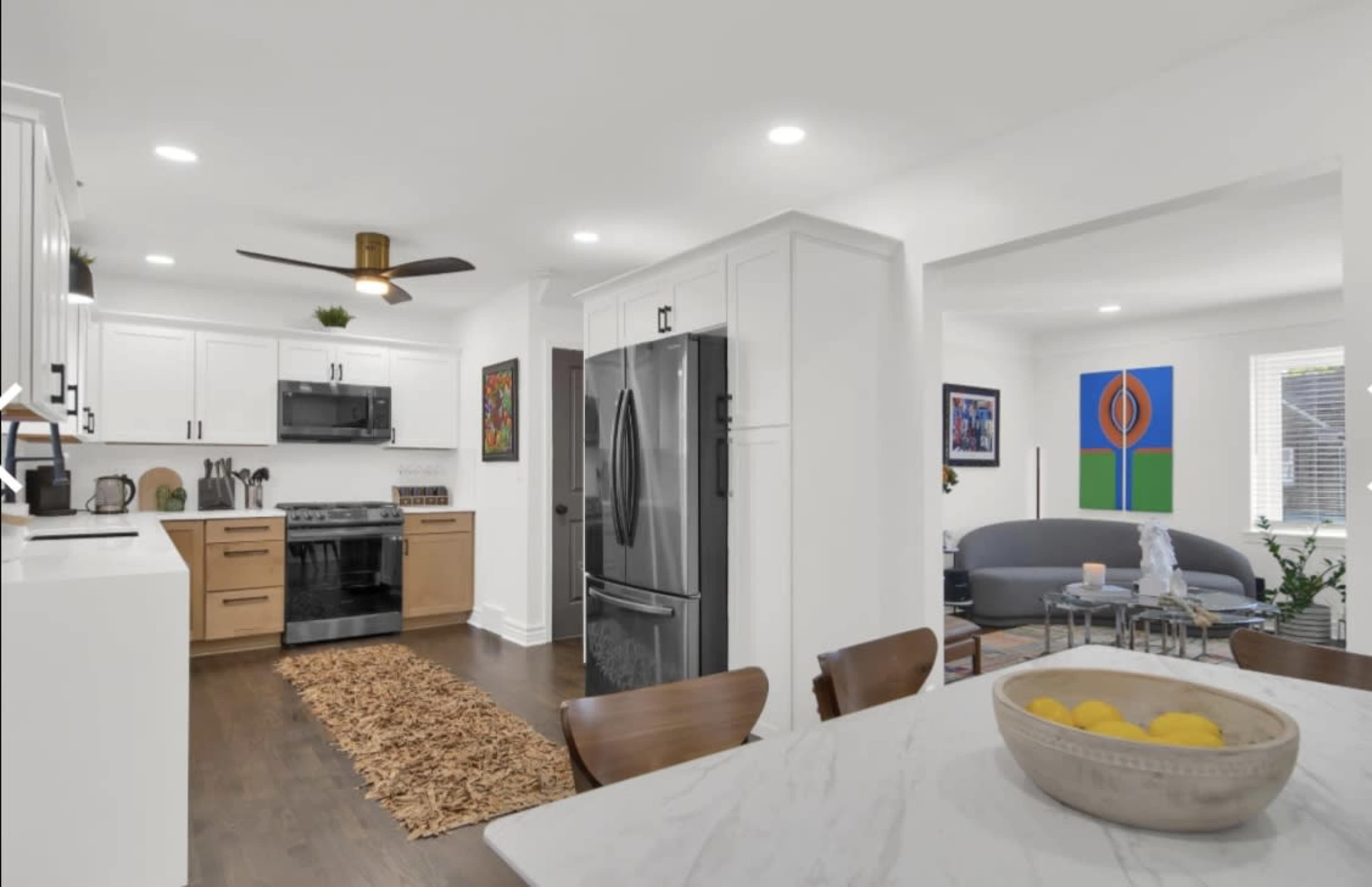 The image shows a modern kitchen and dining area featuring white cabinetry, a dark refrigerator, a gas stove, and a dining table with a bowl of lemons.