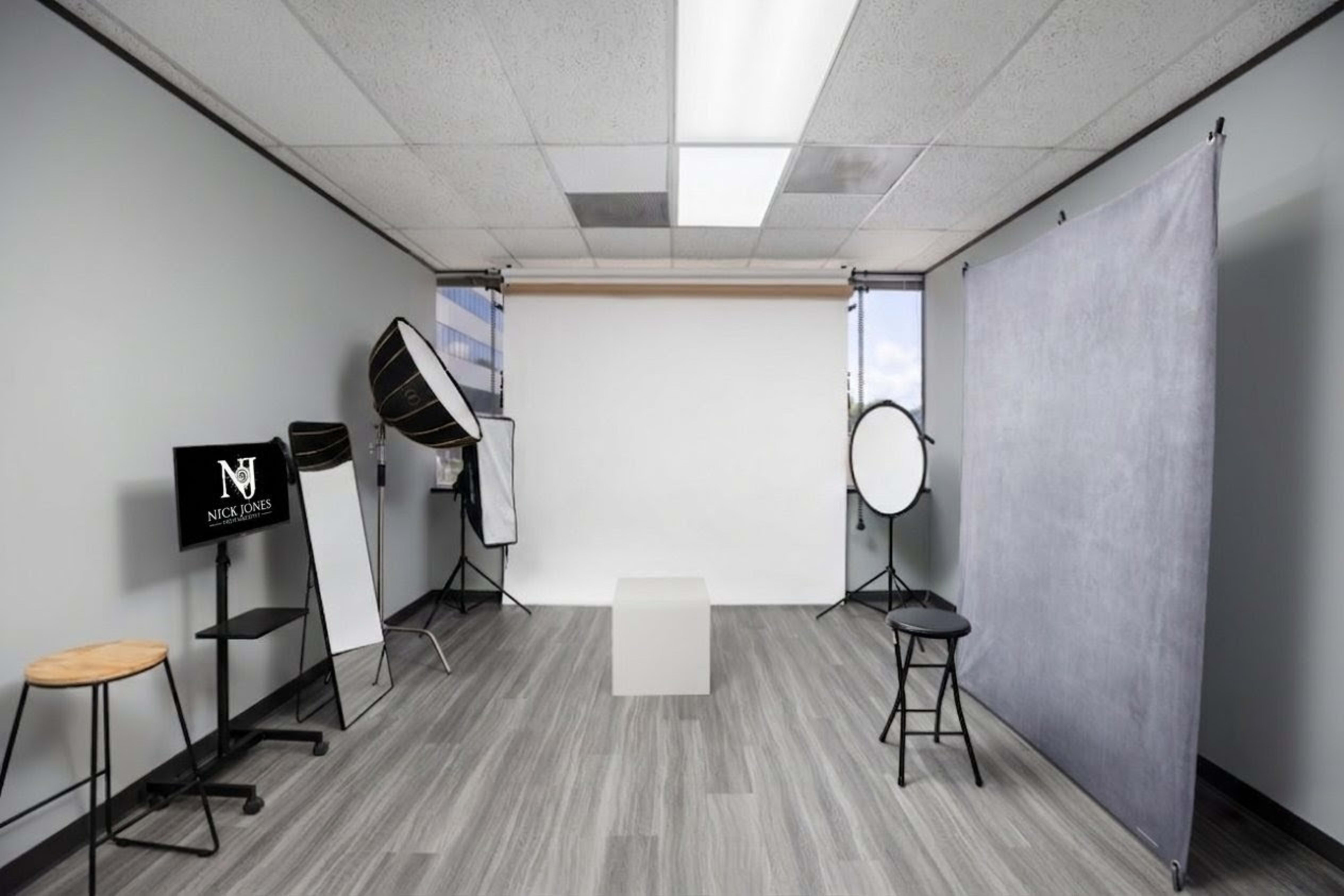 The image shows a photography studio with a white backdrop, various lighting equipment, and seating arrangements for subjects.