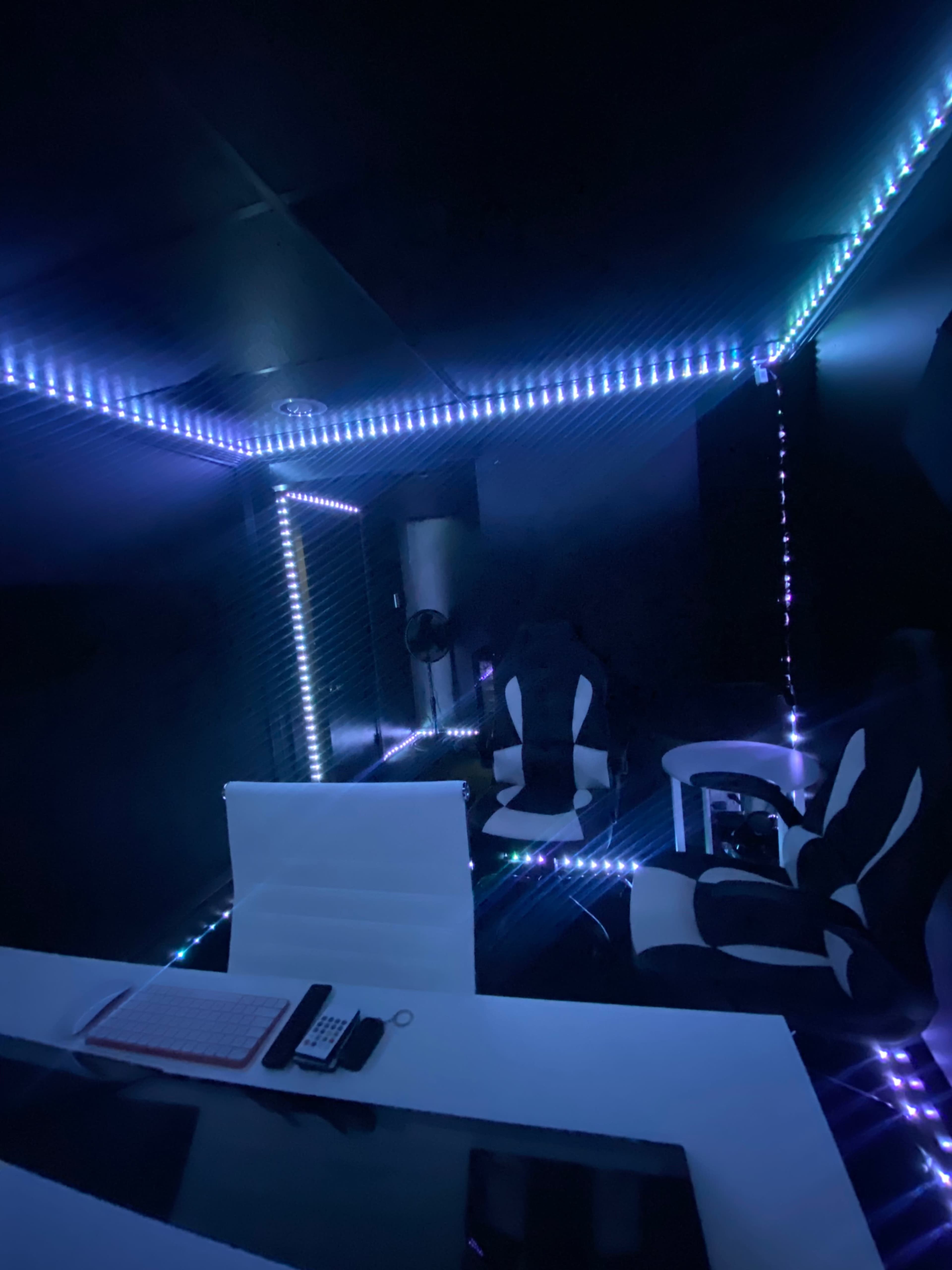 A modern, dimly lit room with black walls and LED lights outlining the corners, featuring a sleek white desk and gaming chairs.