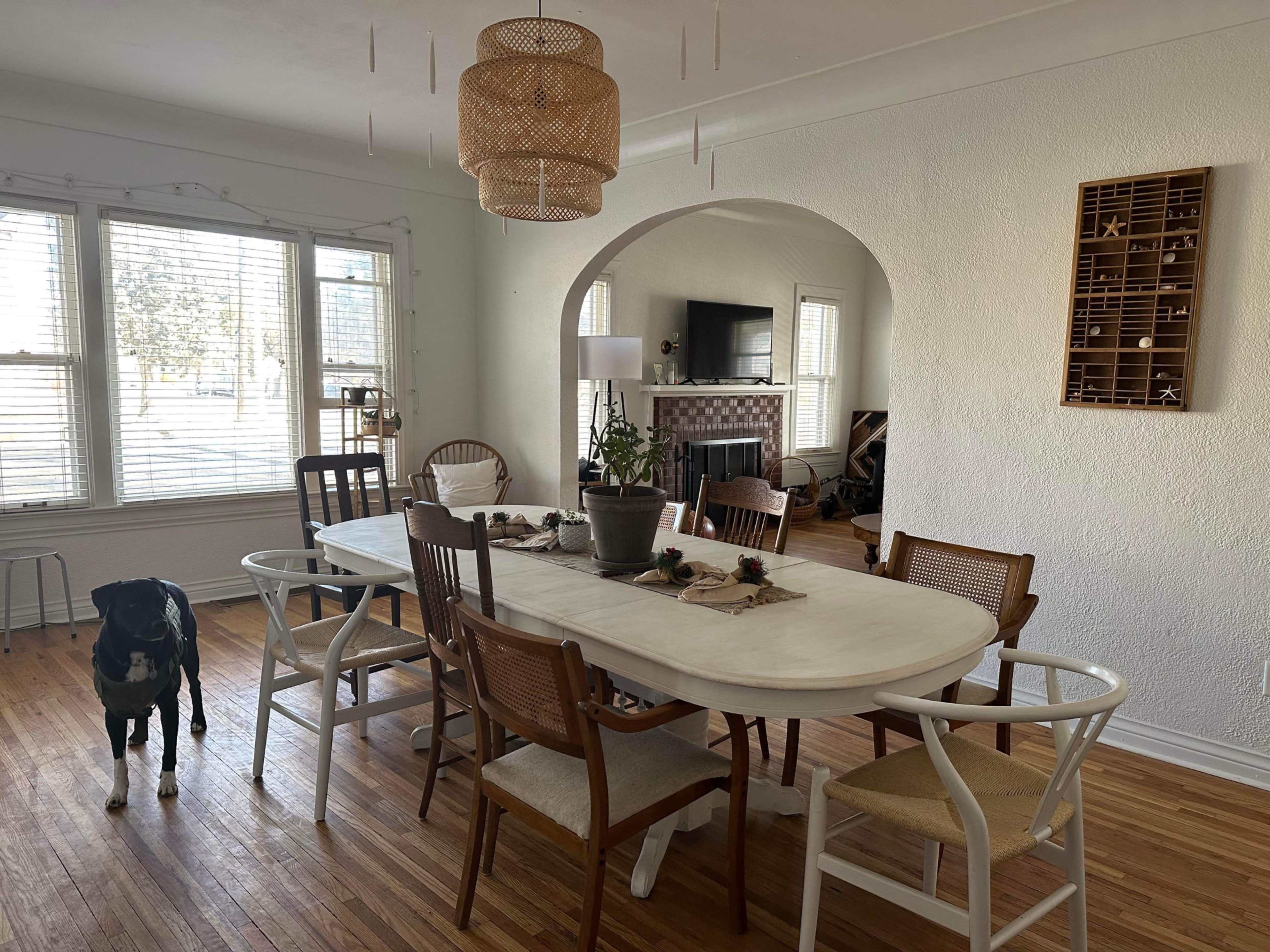 A dining area features a long table surrounded by various chairs, with a large window providing natural light and a living room visible in the background.