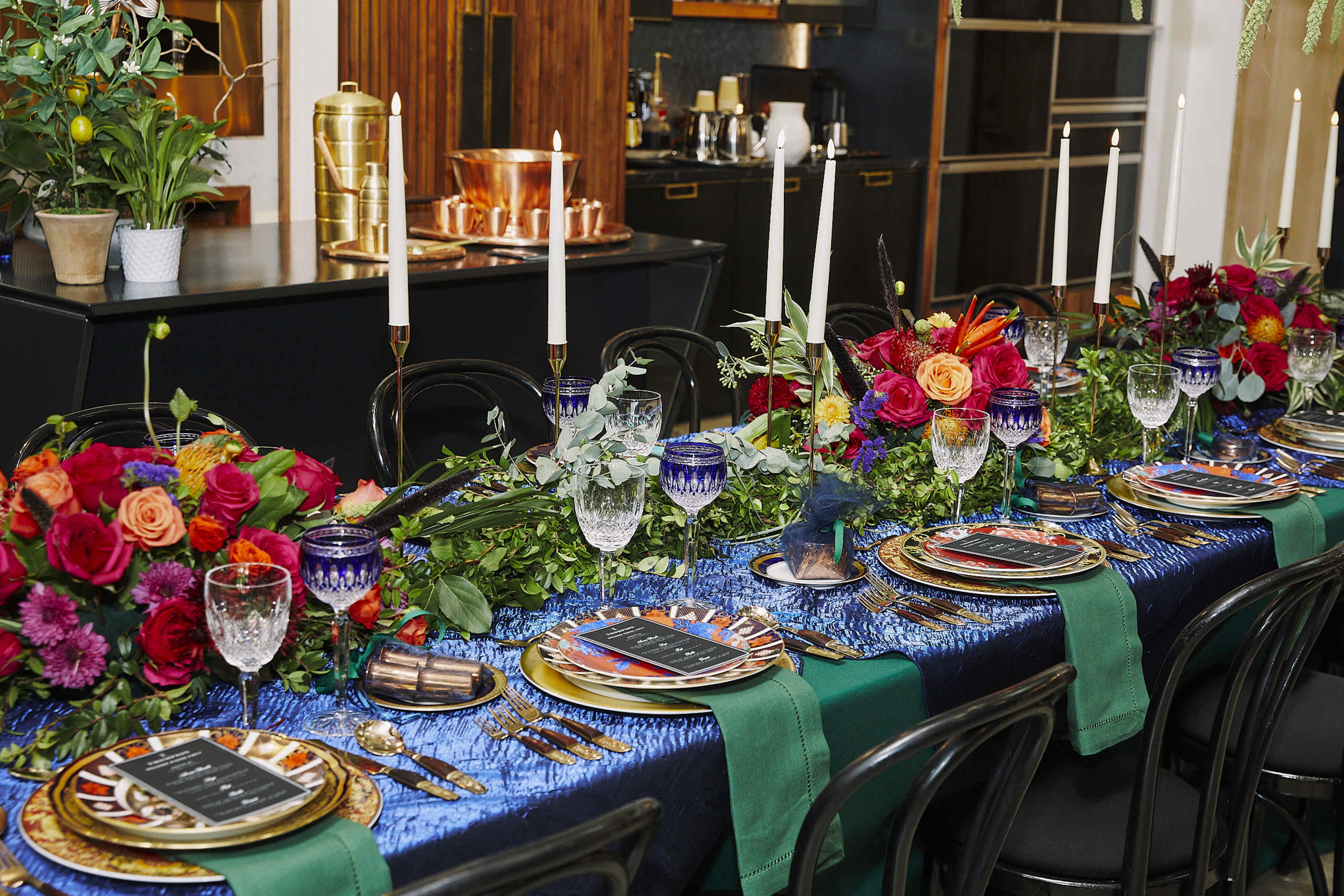A long dining table is beautifully set with colorful floral arrangements, elegant glassware, and decorative plates, surrounded by candles and greenery.