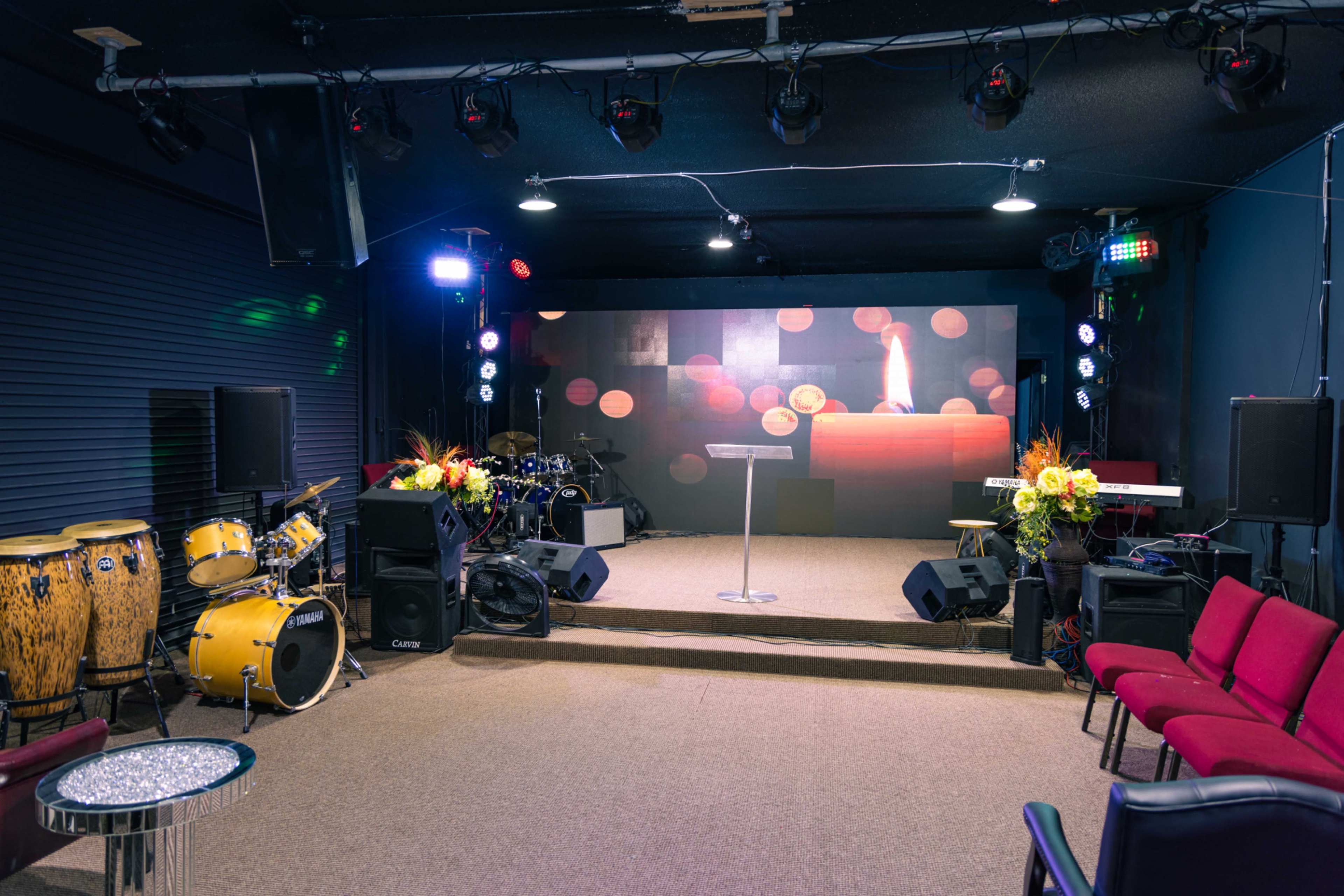 A small stage with a podium and a candle backdrop, surrounded by musical instruments and chairs arranged in front.
