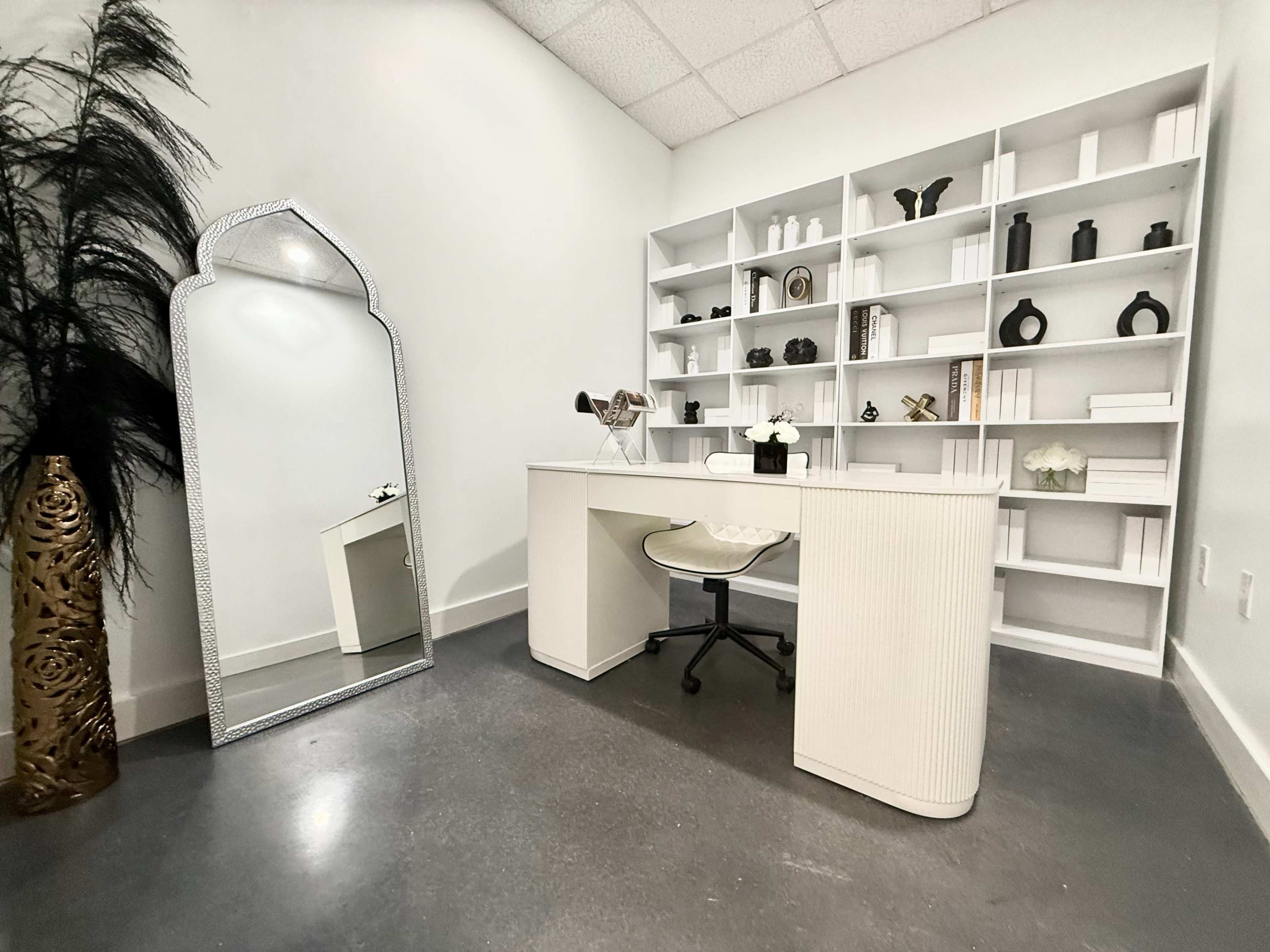 The scene features a minimalist office space with a white desk and a mirrored decorative arch, accompanied by a bookshelf displaying various objects and a decorative plant in the corner.