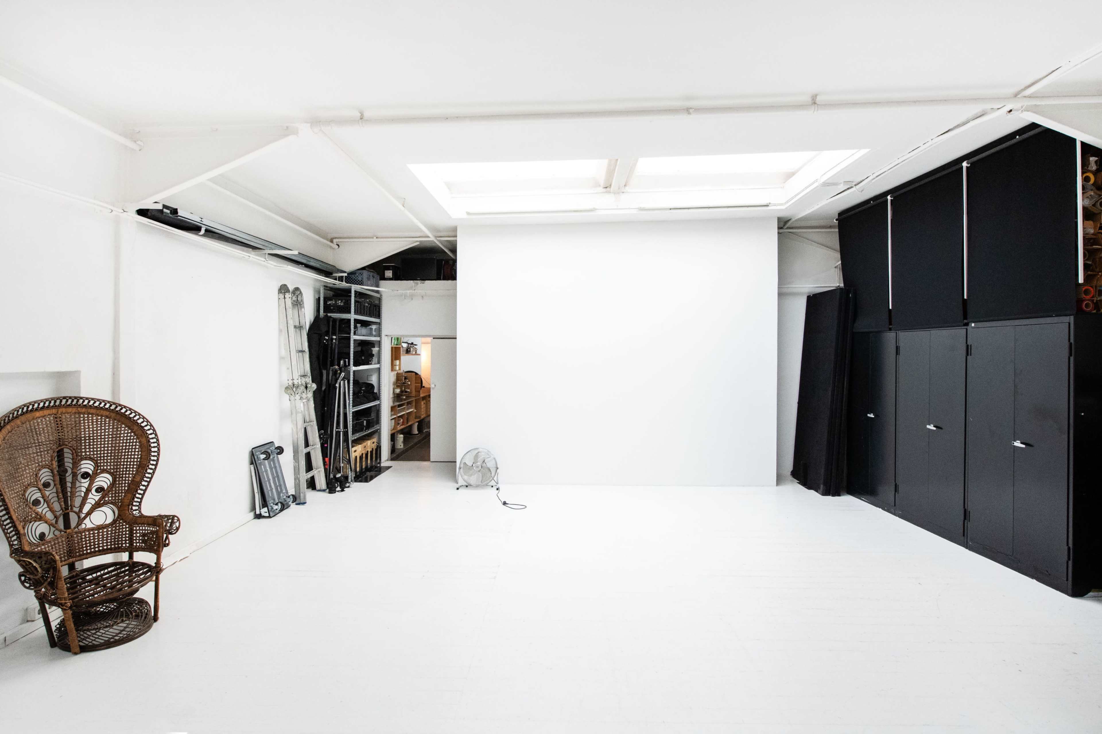 The image shows a spacious, white-walled studio with a large blank wall, a decorative chair, and storage units along one side.