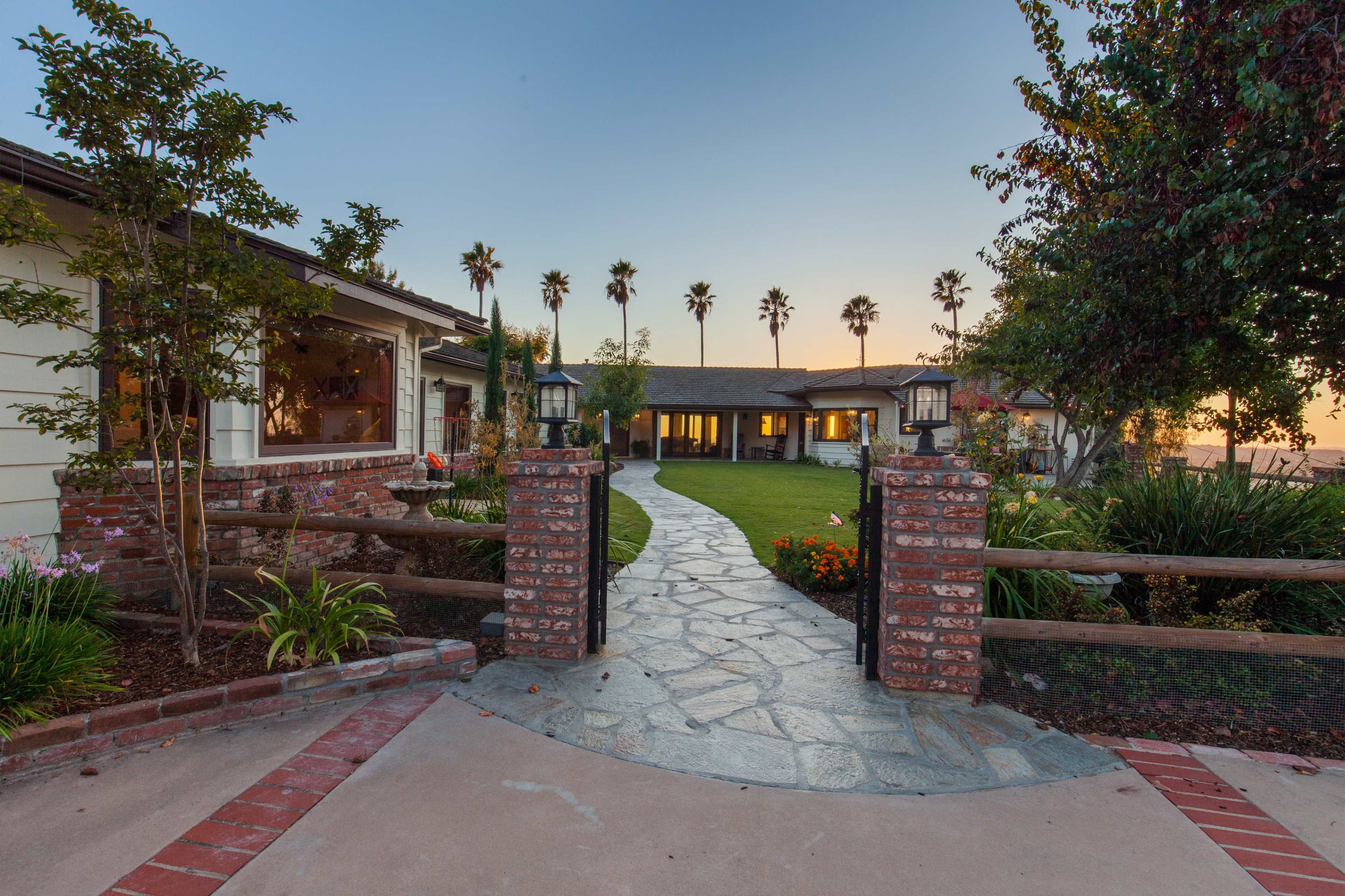 A stone pathway leads through a landscaped entrance to a single-story house surrounded by palm trees at dusk.
