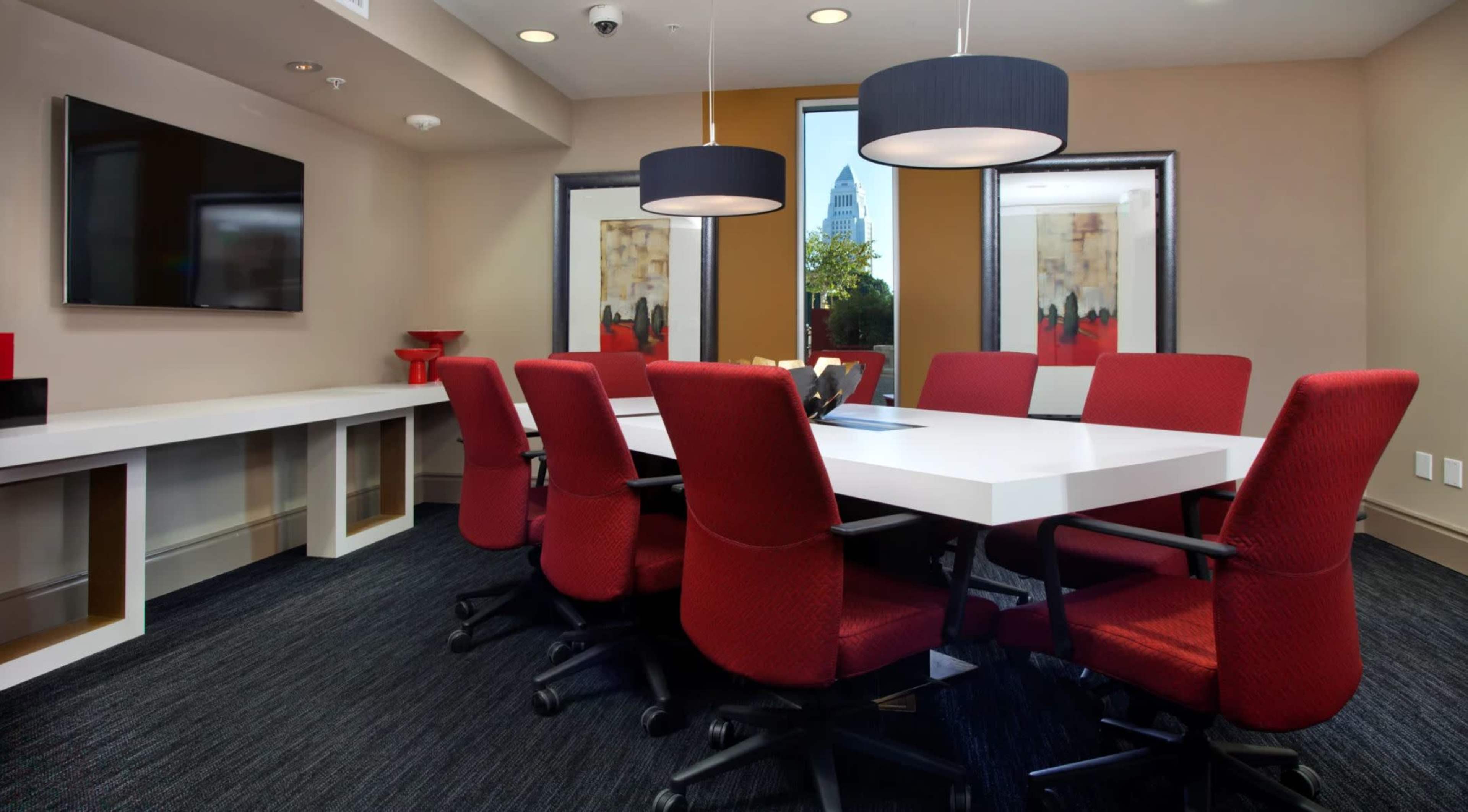 A modern conference room features a large white table surrounded by red chairs, with a TV on the wall and windows showing a city view.