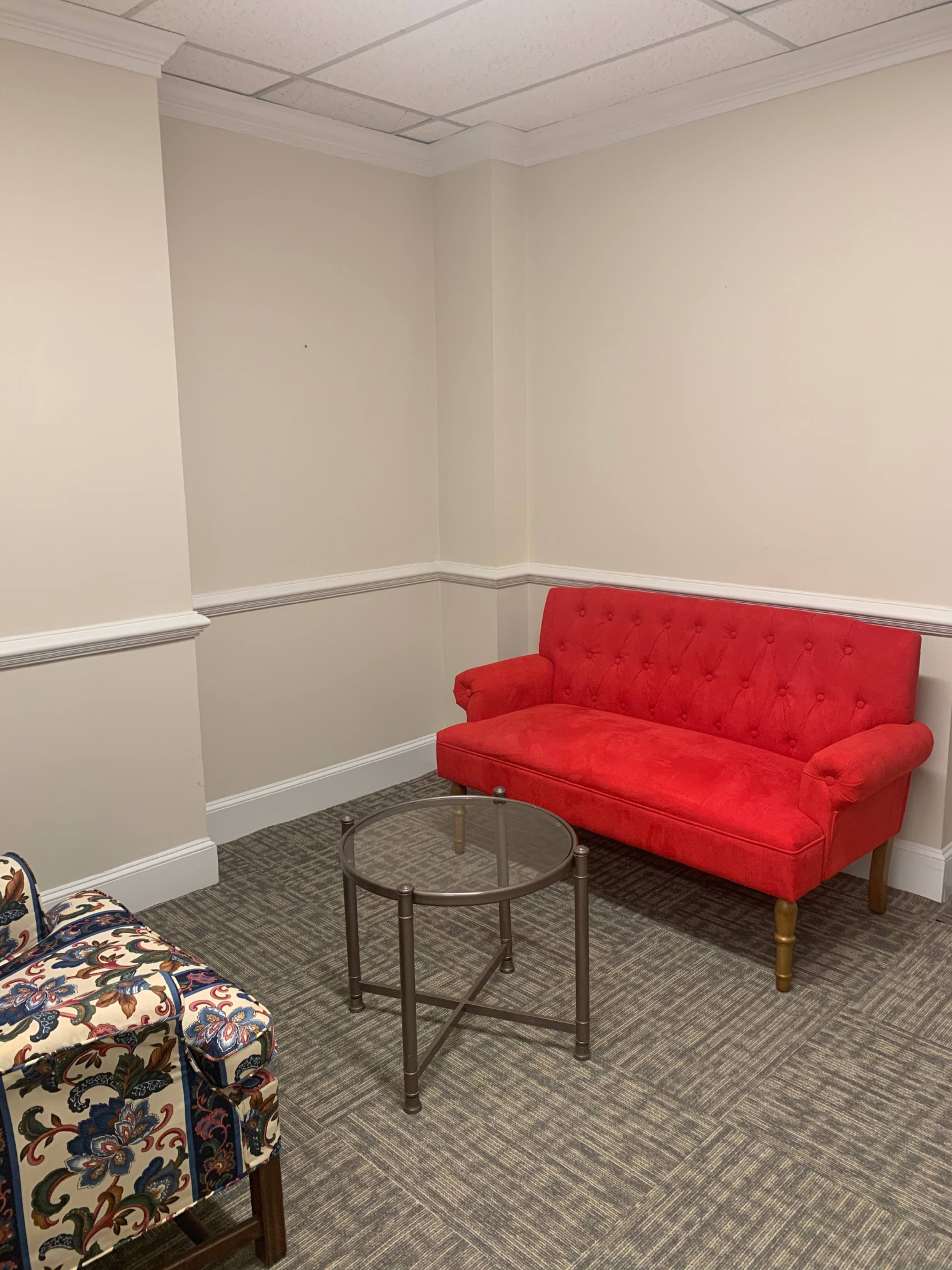 The image shows a small room with a red tufted sofa and a glass coffee table on a patterned carpet.