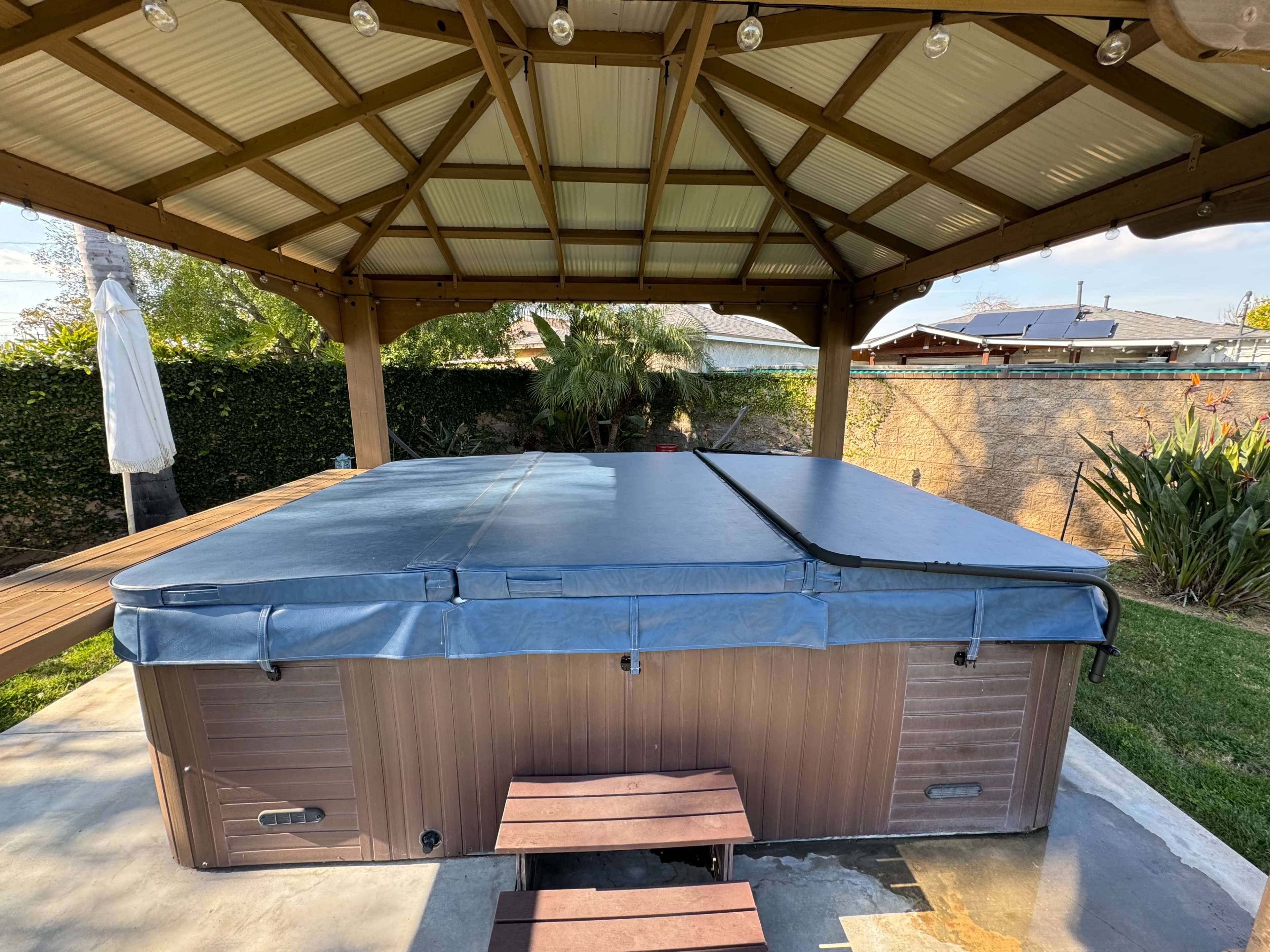 A hot tub with a blue cover is situated under a wooden gazebo in a residential backyard.