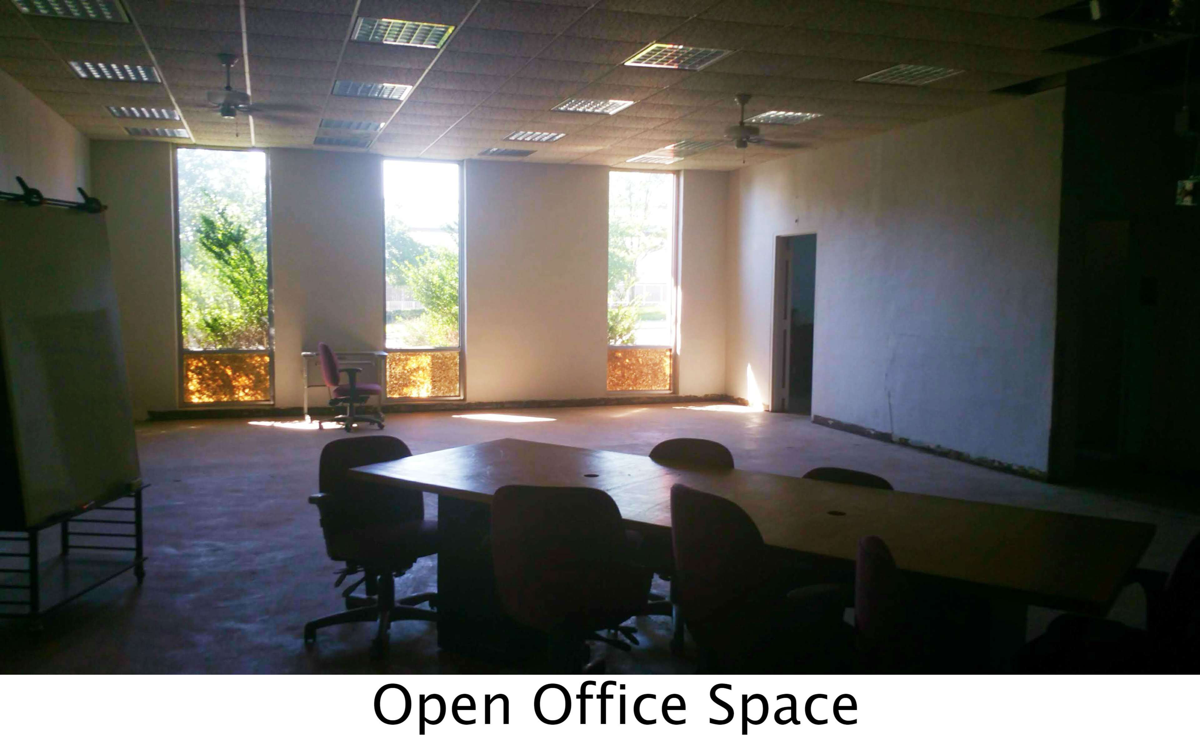 An open office space featuring a large table surrounded by several chairs, with large windows allowing natural light to enter.
