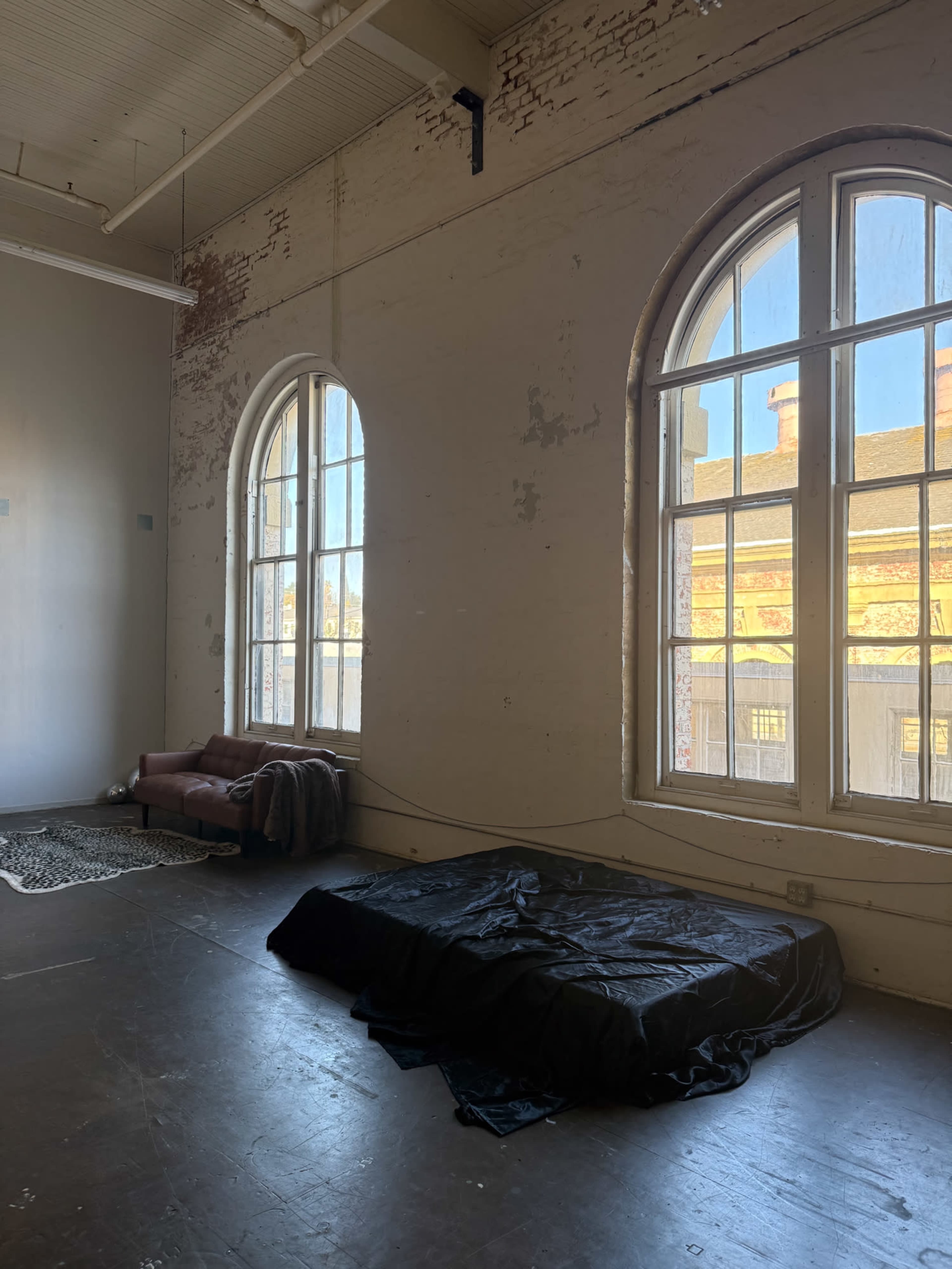 The image shows a spacious room with large arched windows, a couch in the corner, and a bed covered with a black tarp on the floor.