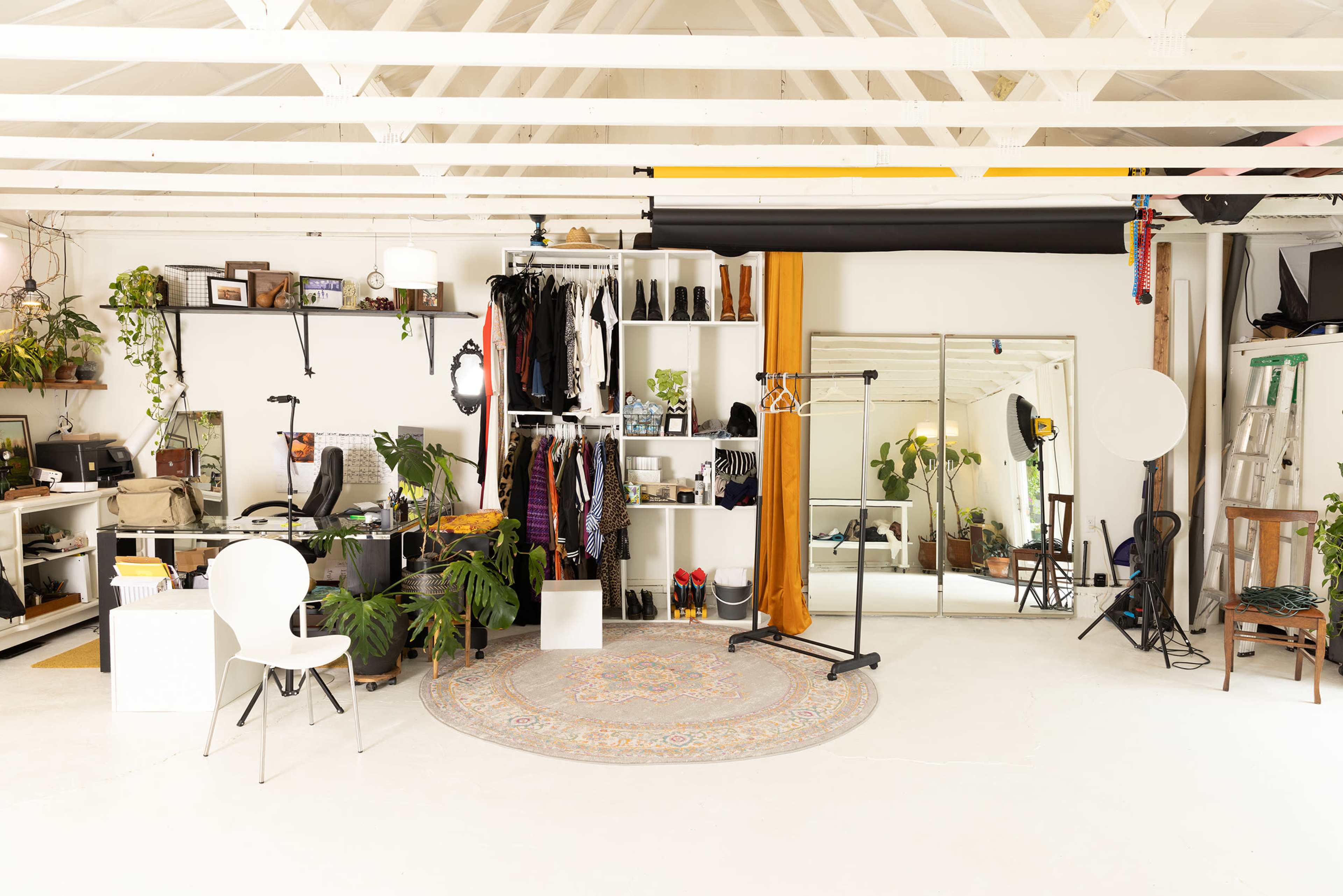 The image shows a brightly lit creative studio space featuring organized shelves with clothing and plants, a circular rug, a mirror, and various equipment scattered throughout.