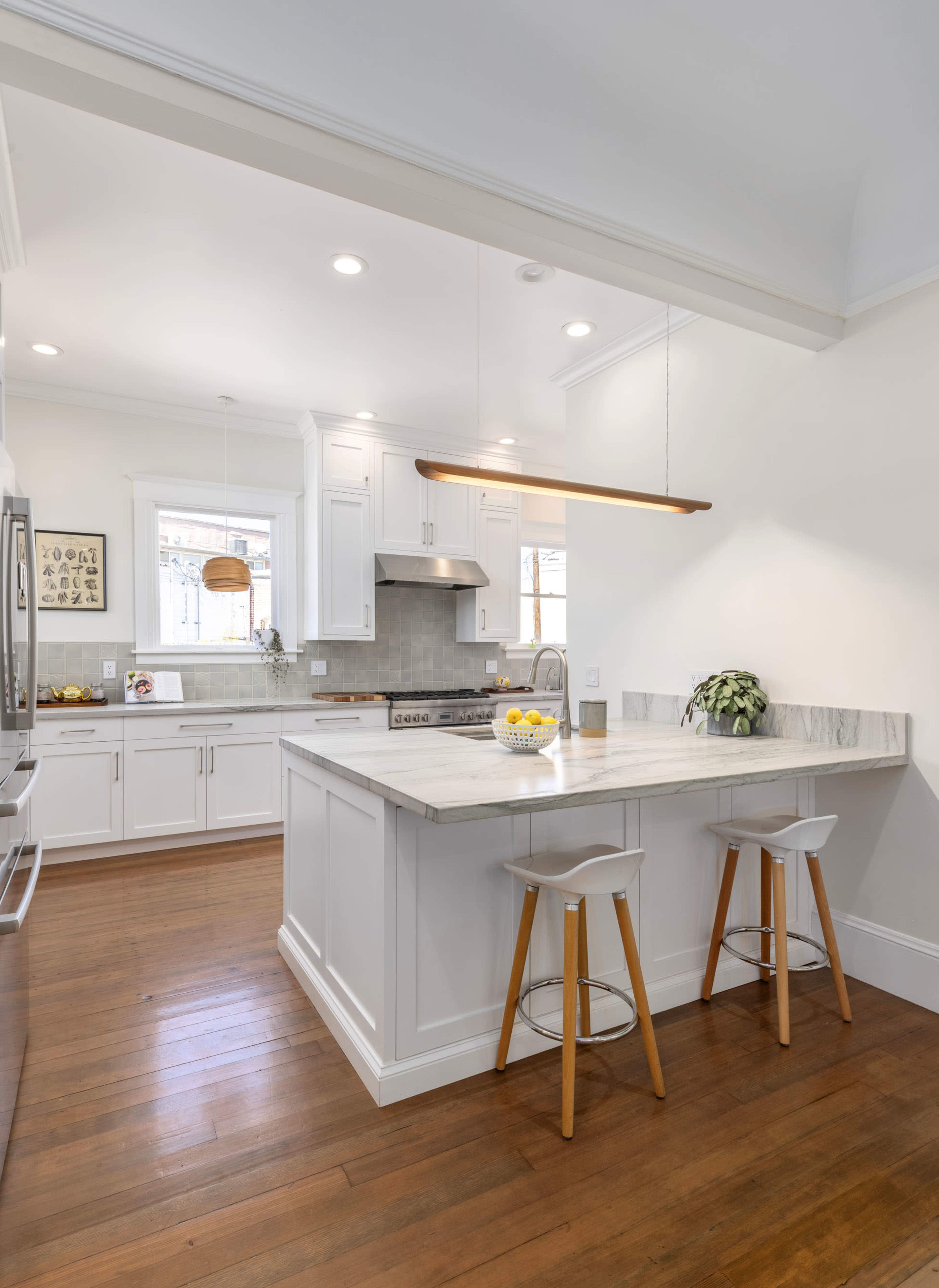 A modern kitchen with white cabinetry, a marble countertop, and two bar stools, illuminated by a pendant light.