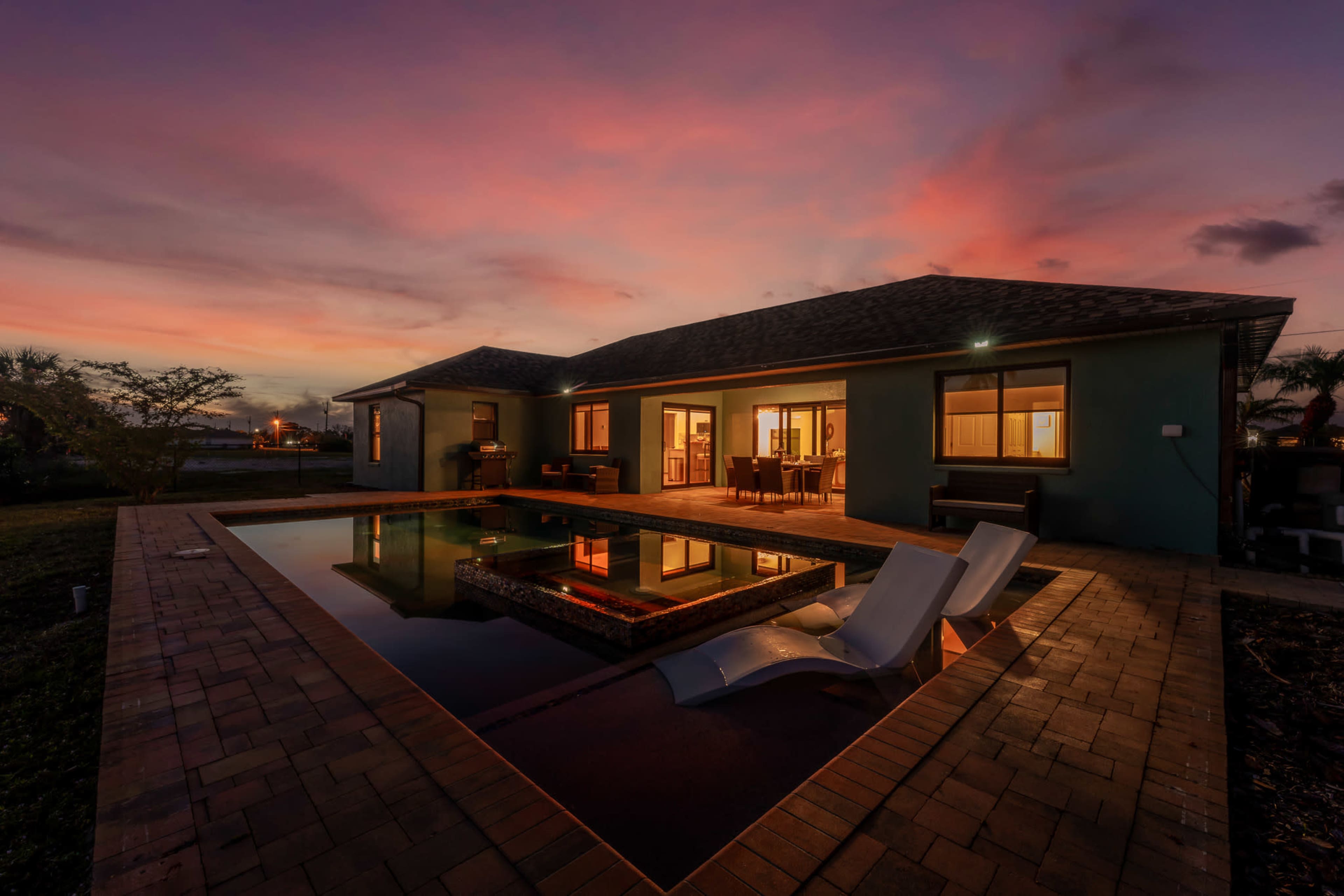 A modern house with a swimming pool reflects a colorful sunset sky in the background.
