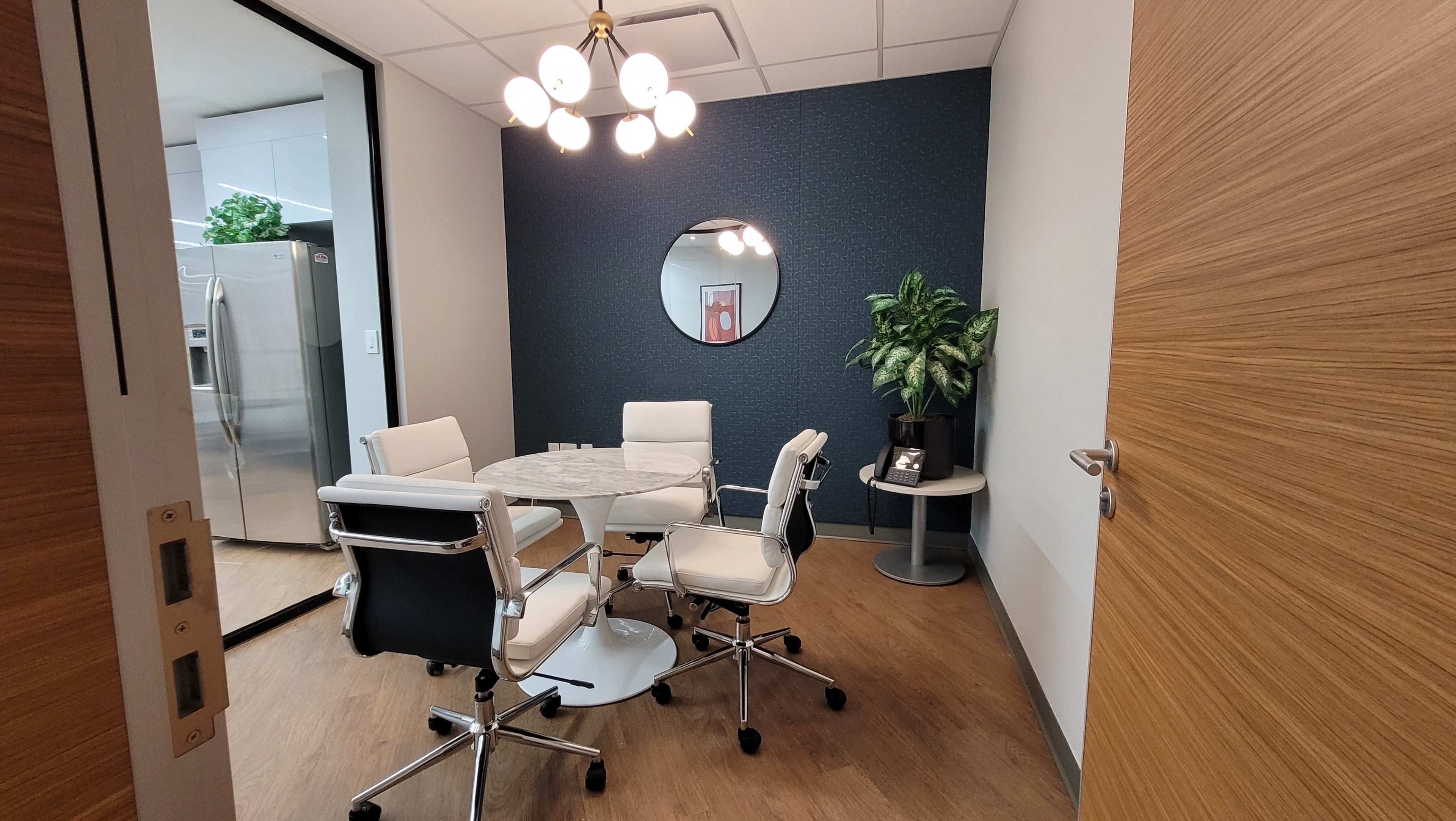 A small meeting room with a round table, four white chairs, a potted plant, and a large mirror on the wall.