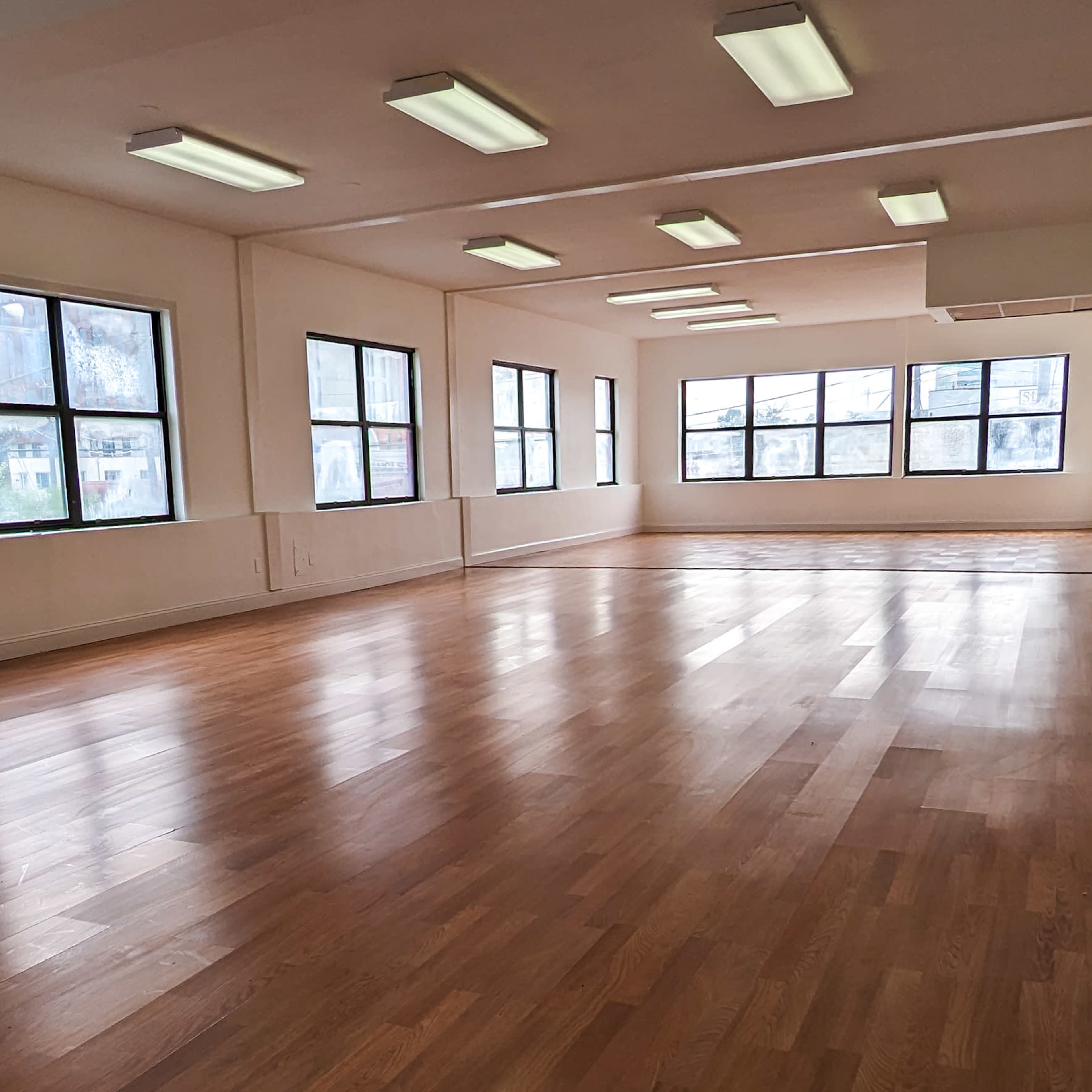 A spacious room with large windows and wooden flooring, illuminated by overhead fluorescent lights.