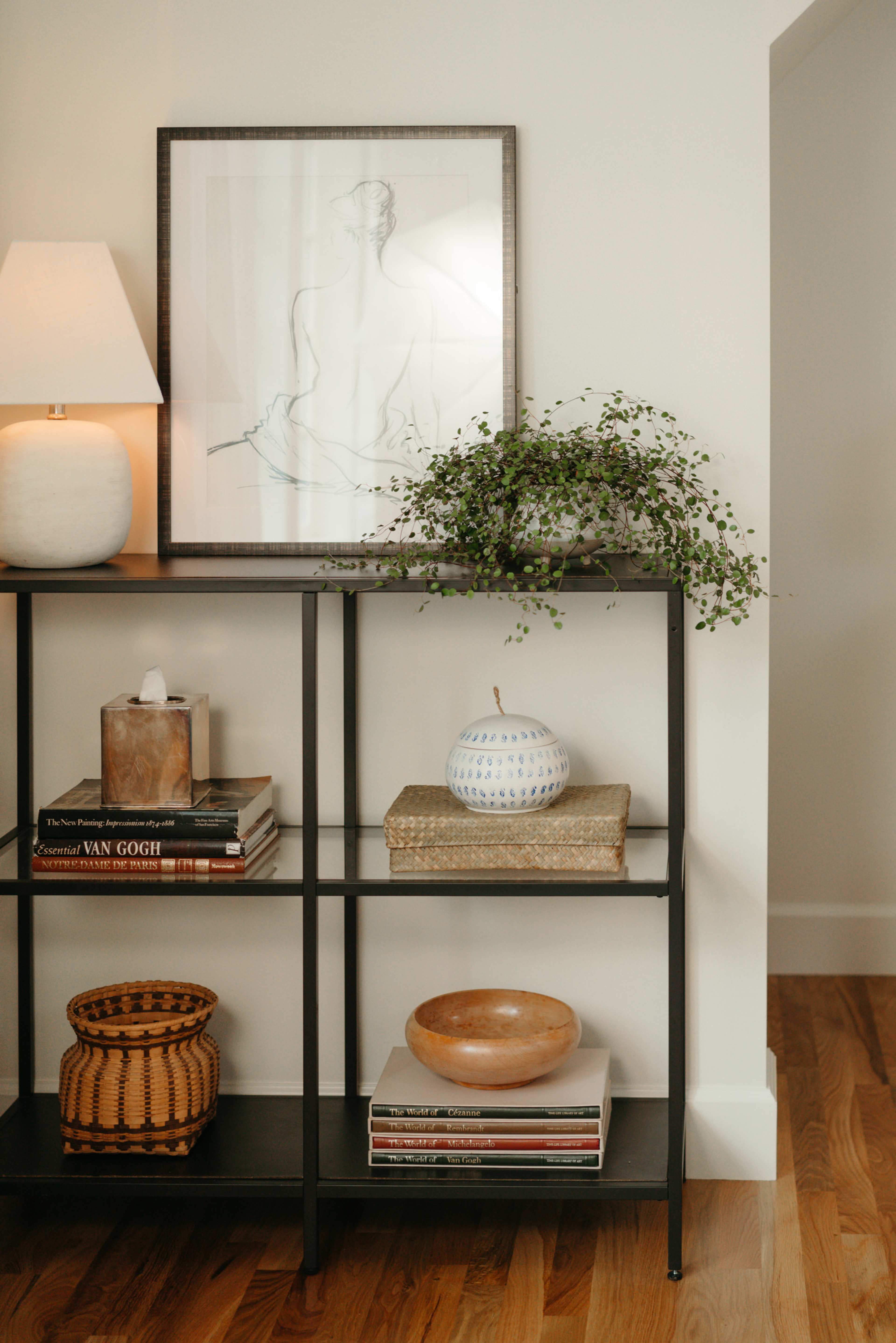 A modern bookshelf displays a framed line drawing, various books, decorative bowls, and a woven basket alongside a lamp and a trailing plant.