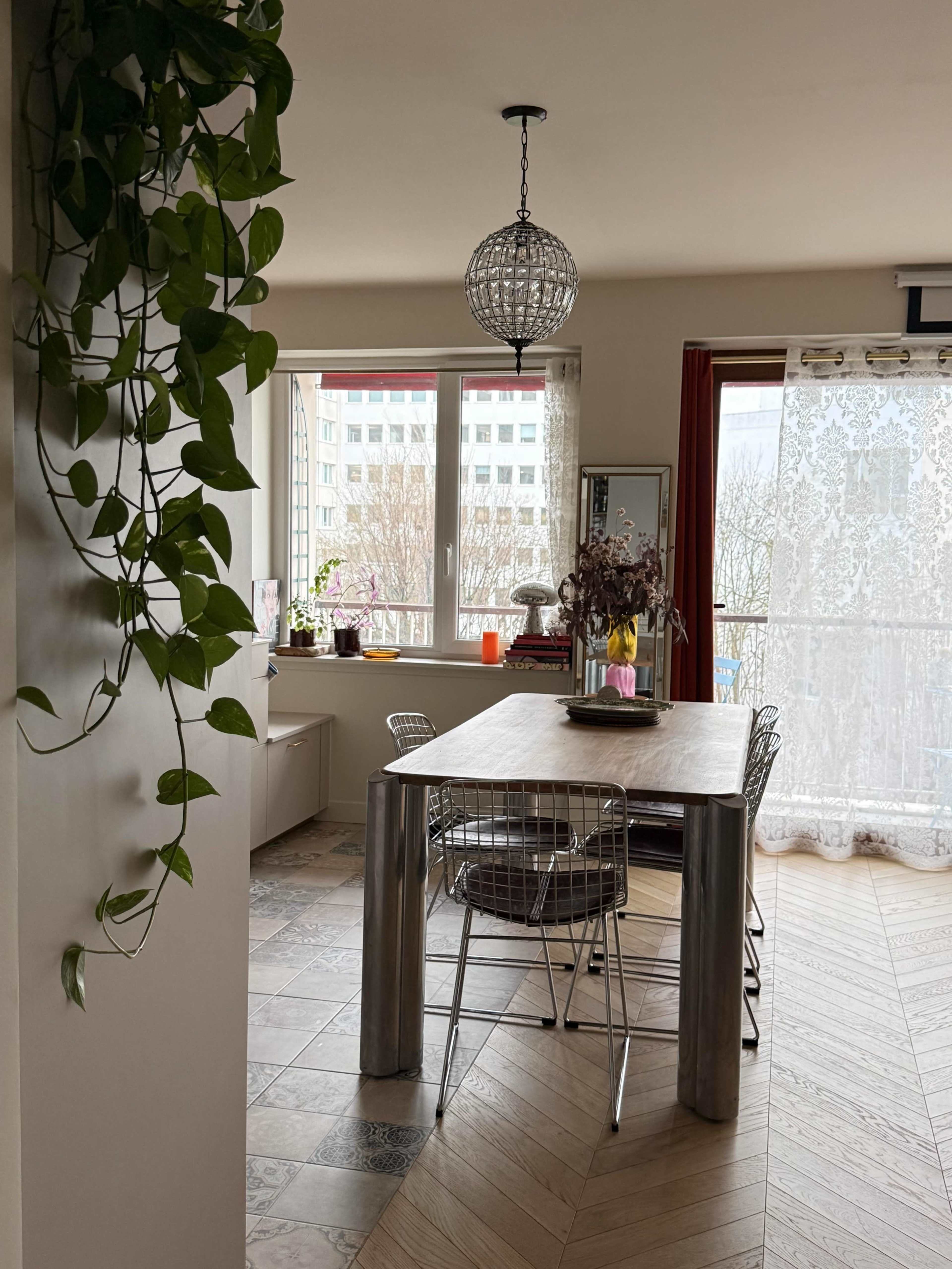 The image shows a dining area featuring a wooden table surrounded by metal chairs, with large windows that allow natural light to illuminate the space.