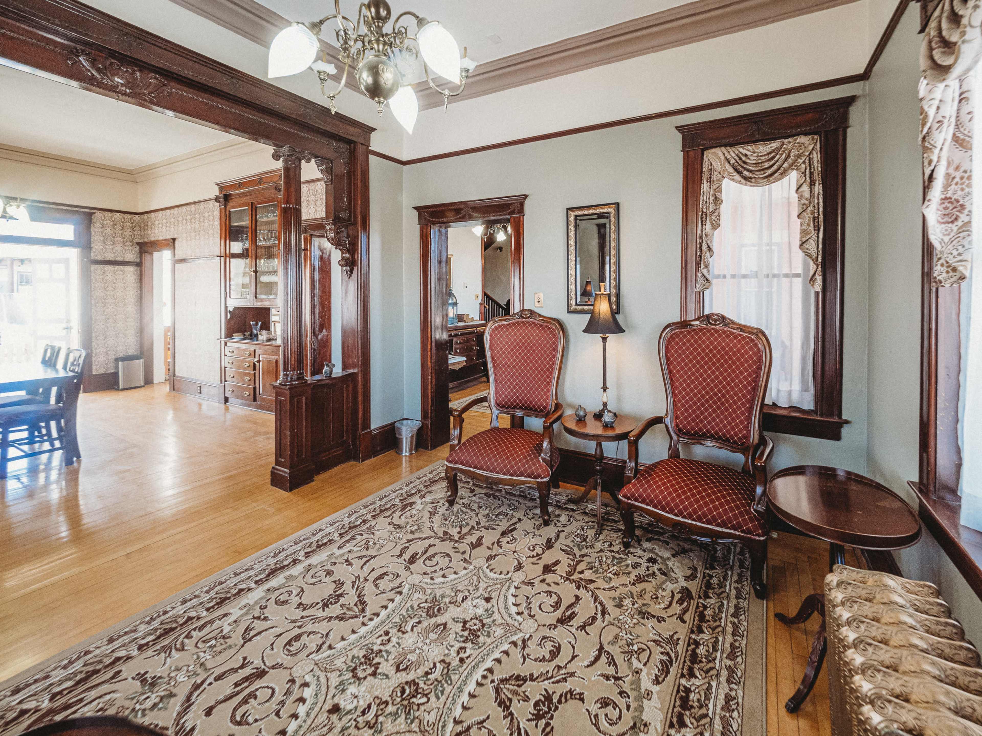 A vintage living room with ornate chairs, a patterned rug, and wooden accents, leading into a dining area.