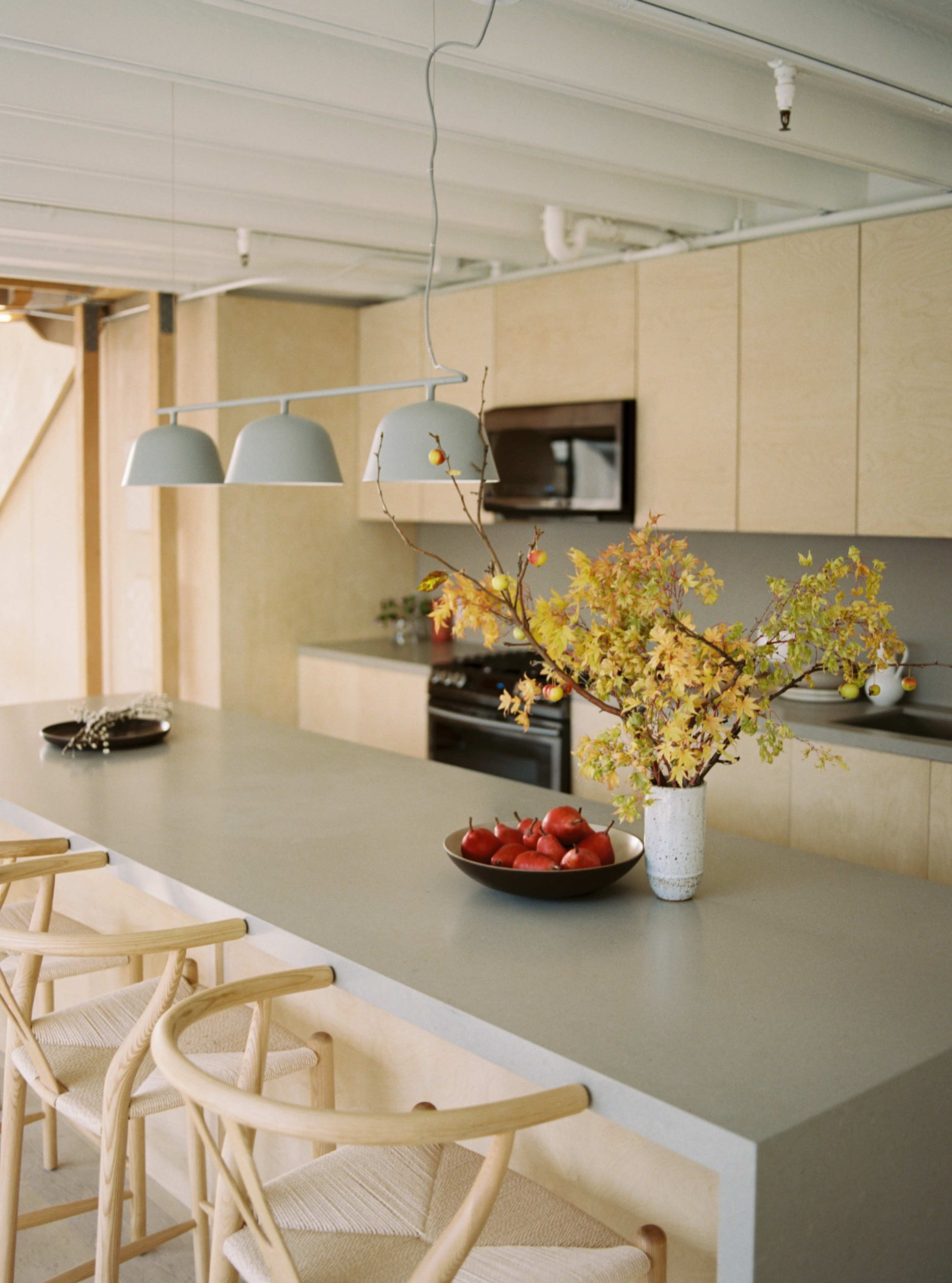 A modern kitchen features a long countertop with bar stools, pendant lights overhead, and a vase of autumn foliage and fruit on the table.