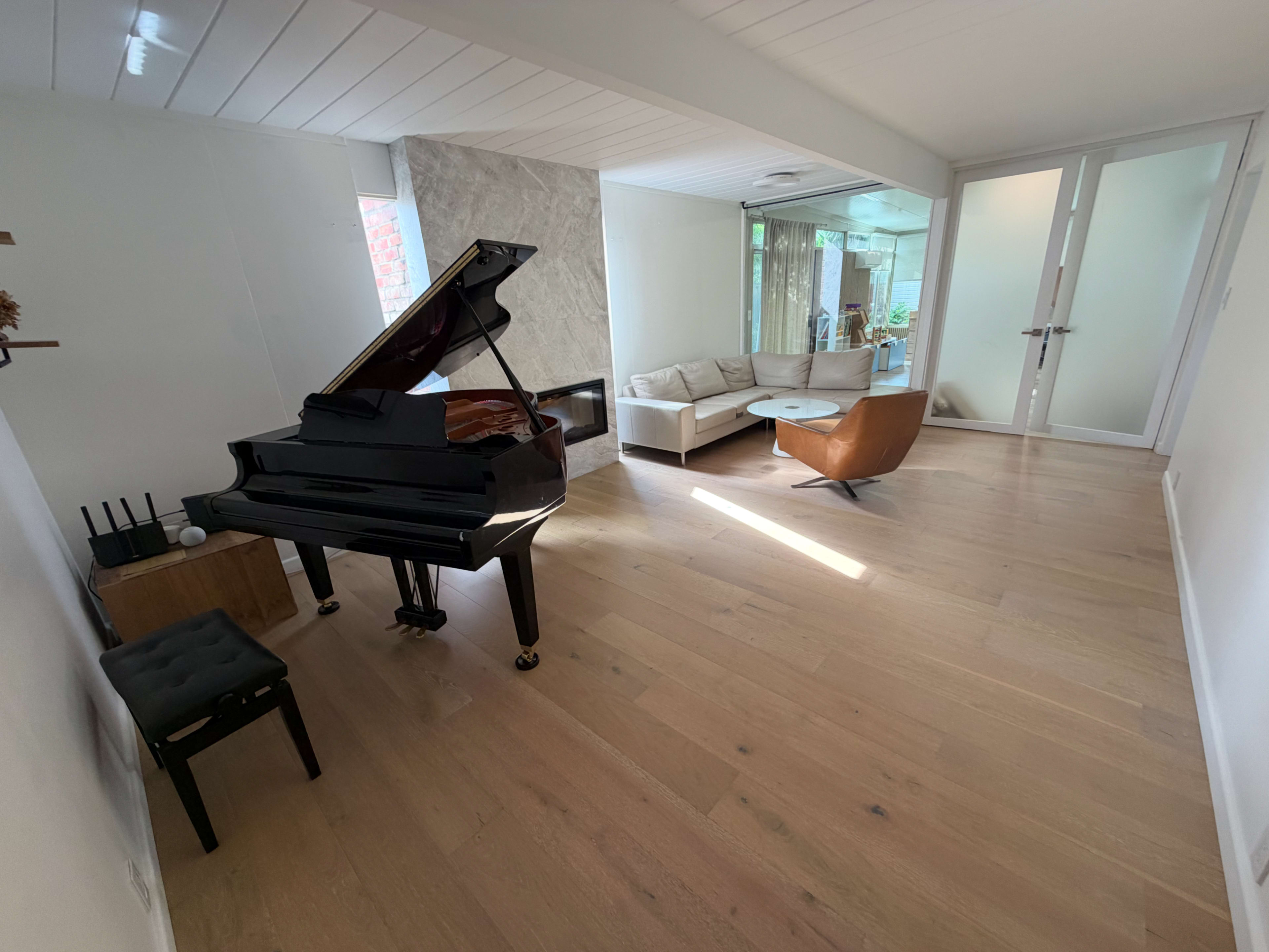 The image shows a spacious, modern living area featuring a grand piano, a small black stool, a light-colored sofa set, and a coffee table, with large windows allowing natural light.