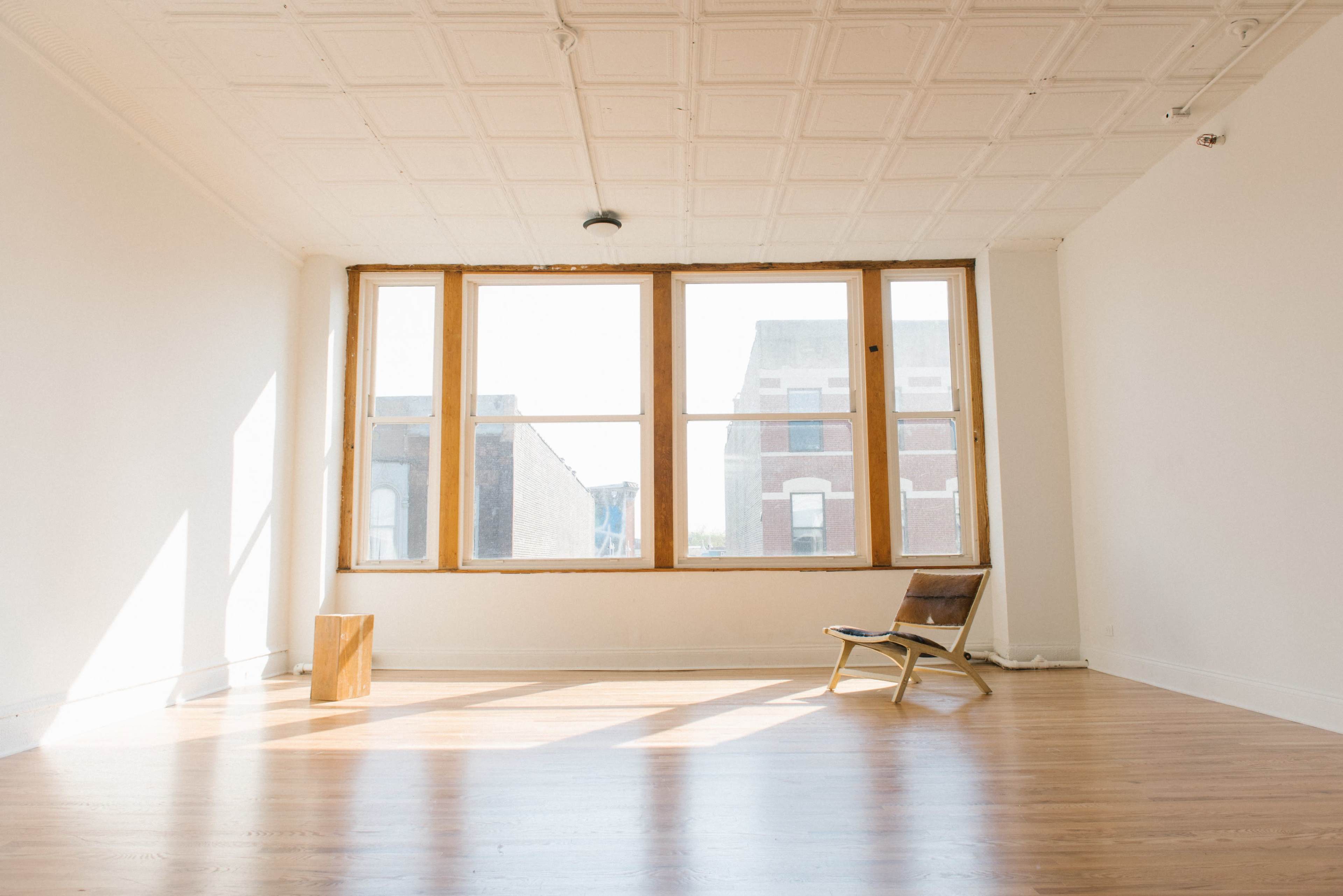 A bright, empty room features a large window with natural light, a wooden chair, and wooden flooring.