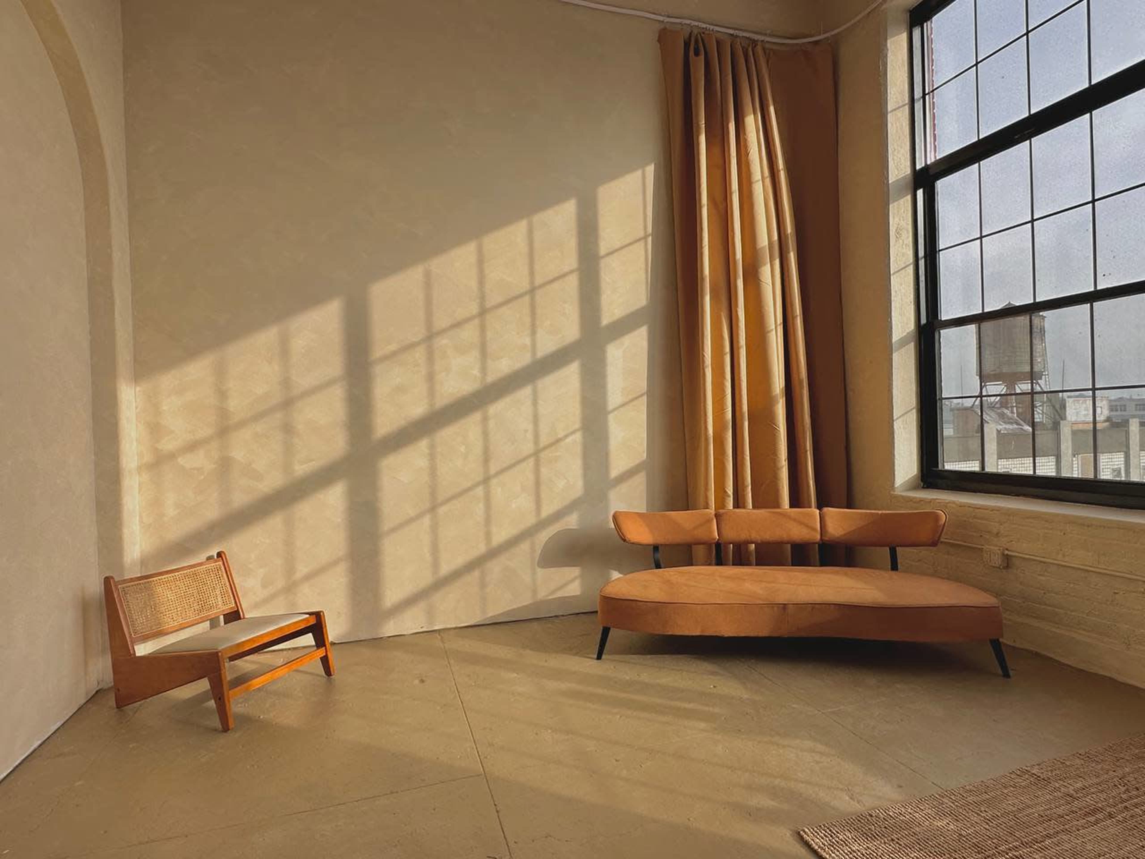 A minimalist interior with a brown couch, a small wooden chair, and large windows casting geometric shadows on the floor.