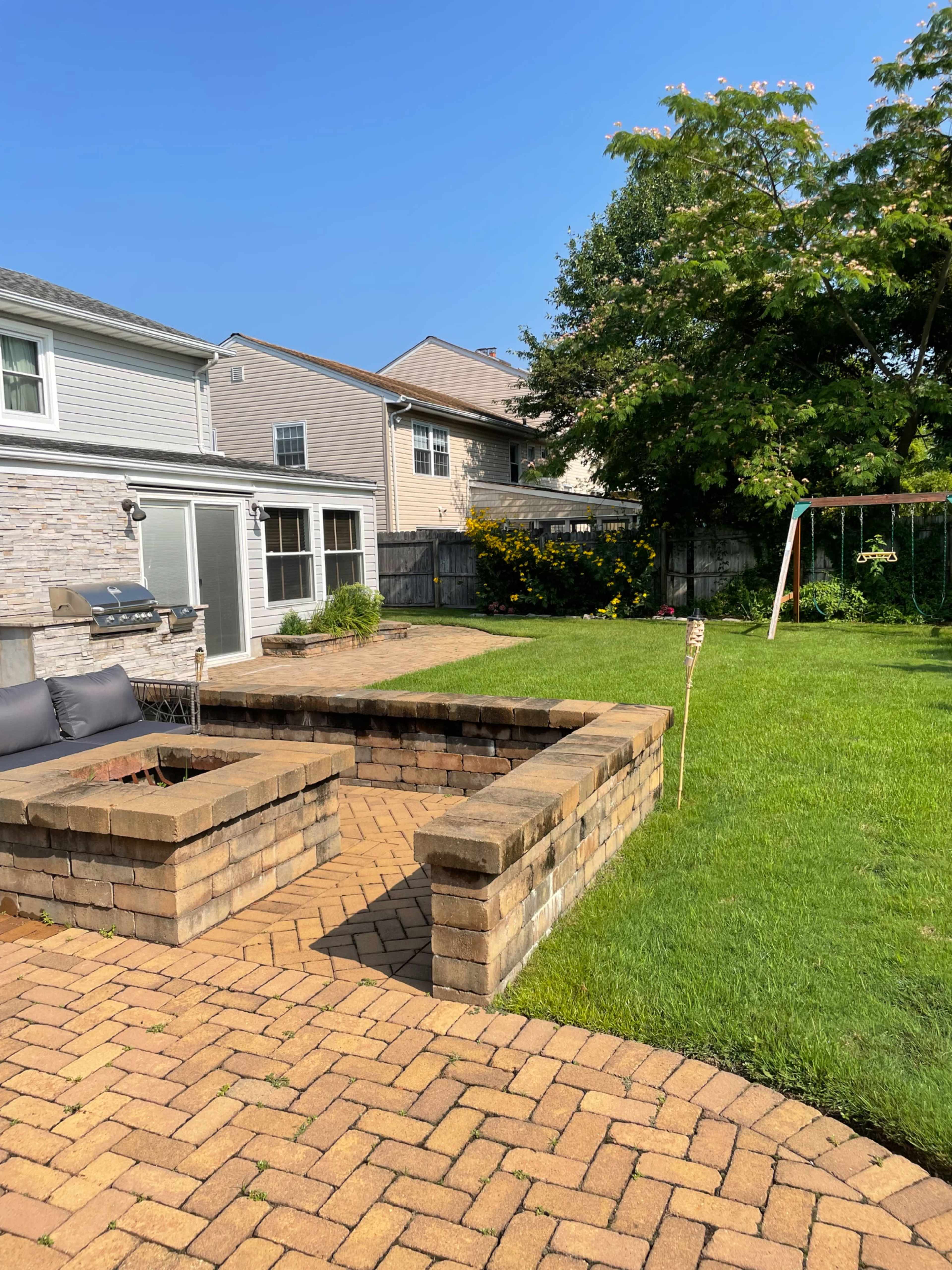A landscaped backyard featuring a stone patio, a fire pit area, a swing set, and lush green grass.