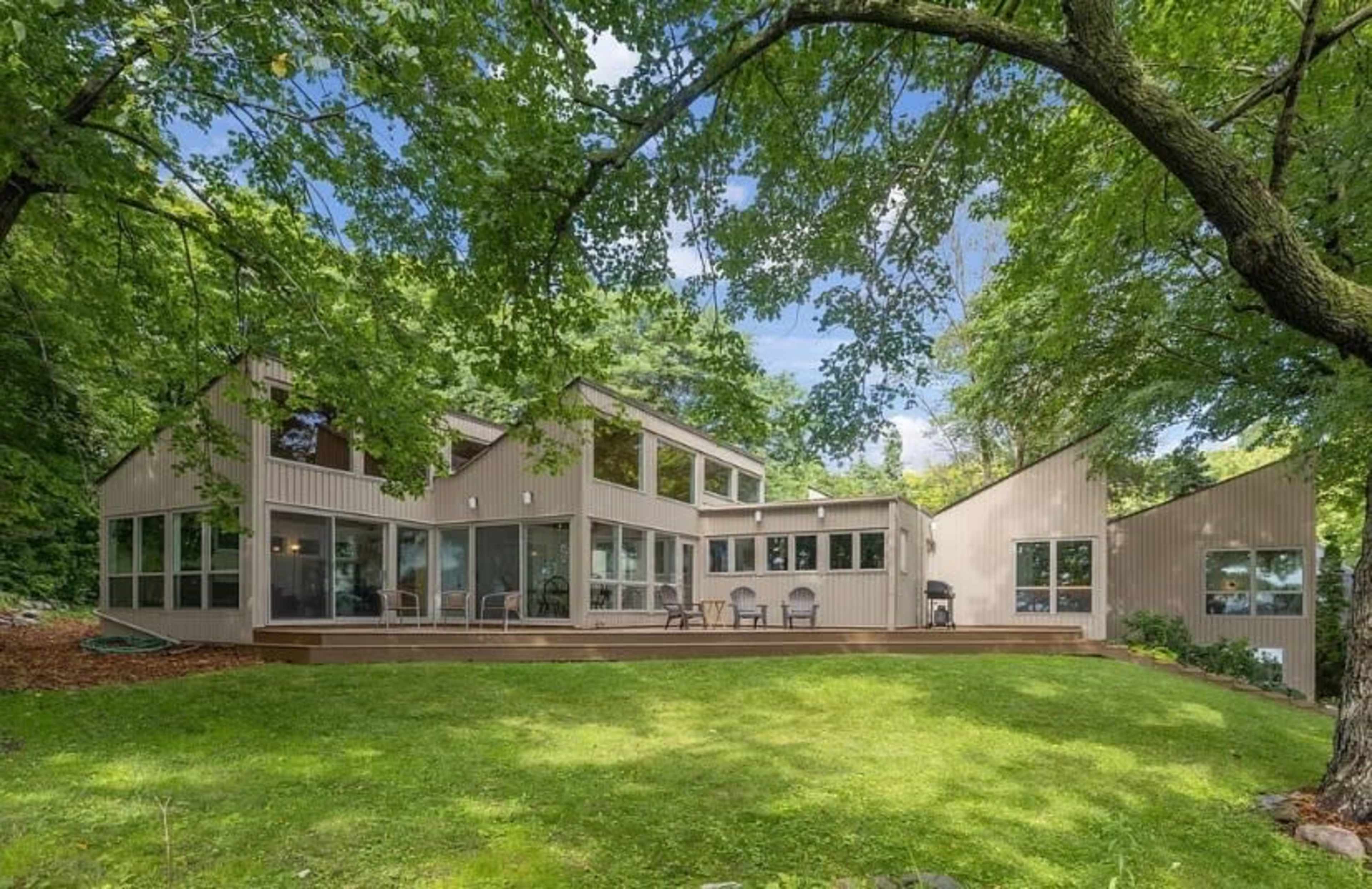 The image shows a modern house surrounded by large trees and a green lawn, featuring large windows and a wooden deck.