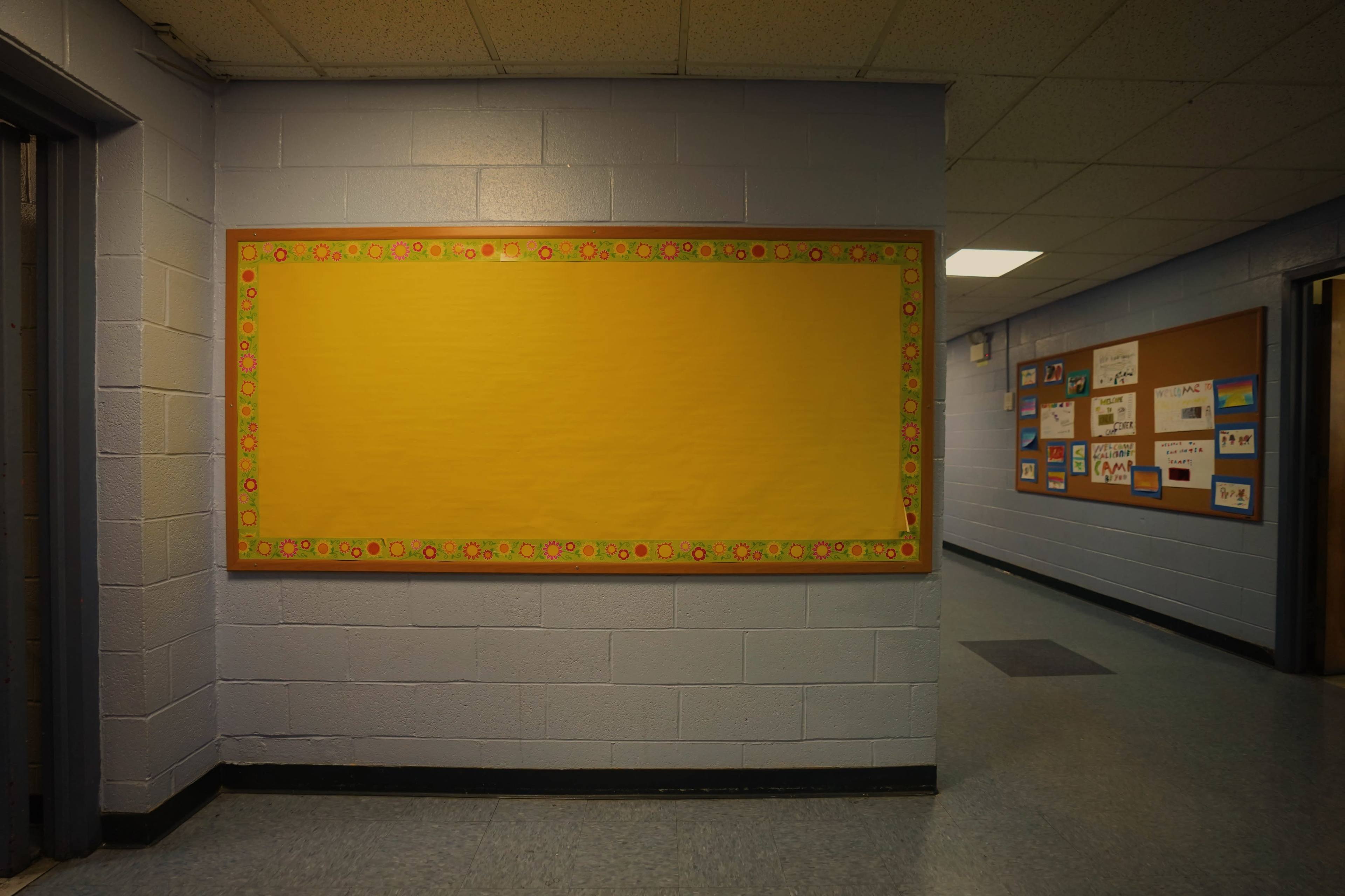 A hallway with a blank, yellow bulletin board on one wall and a display of colorful posters on the opposite wall.