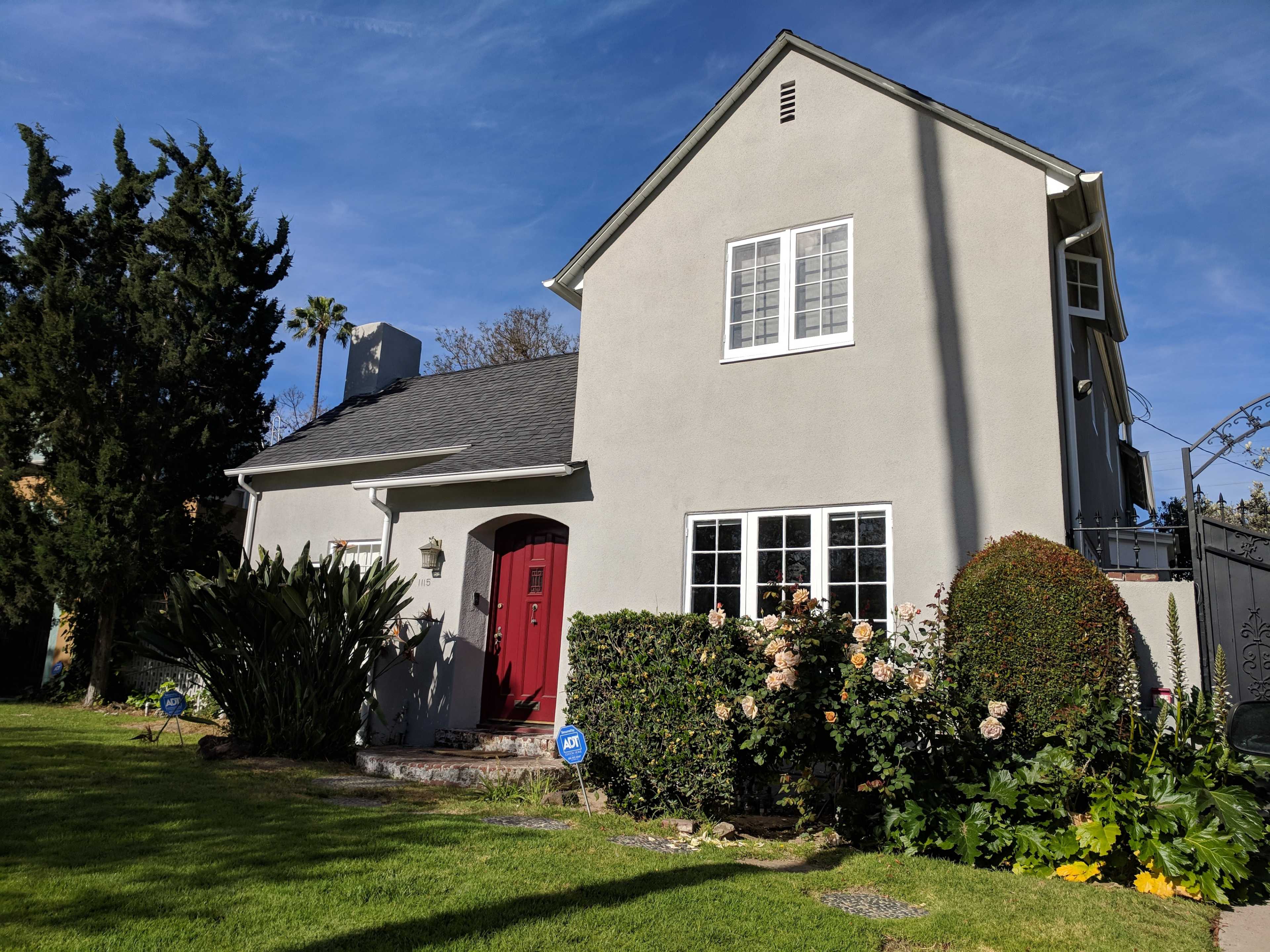 A two-story gray house with a red door is surrounded by greenery and flowers in the garden.