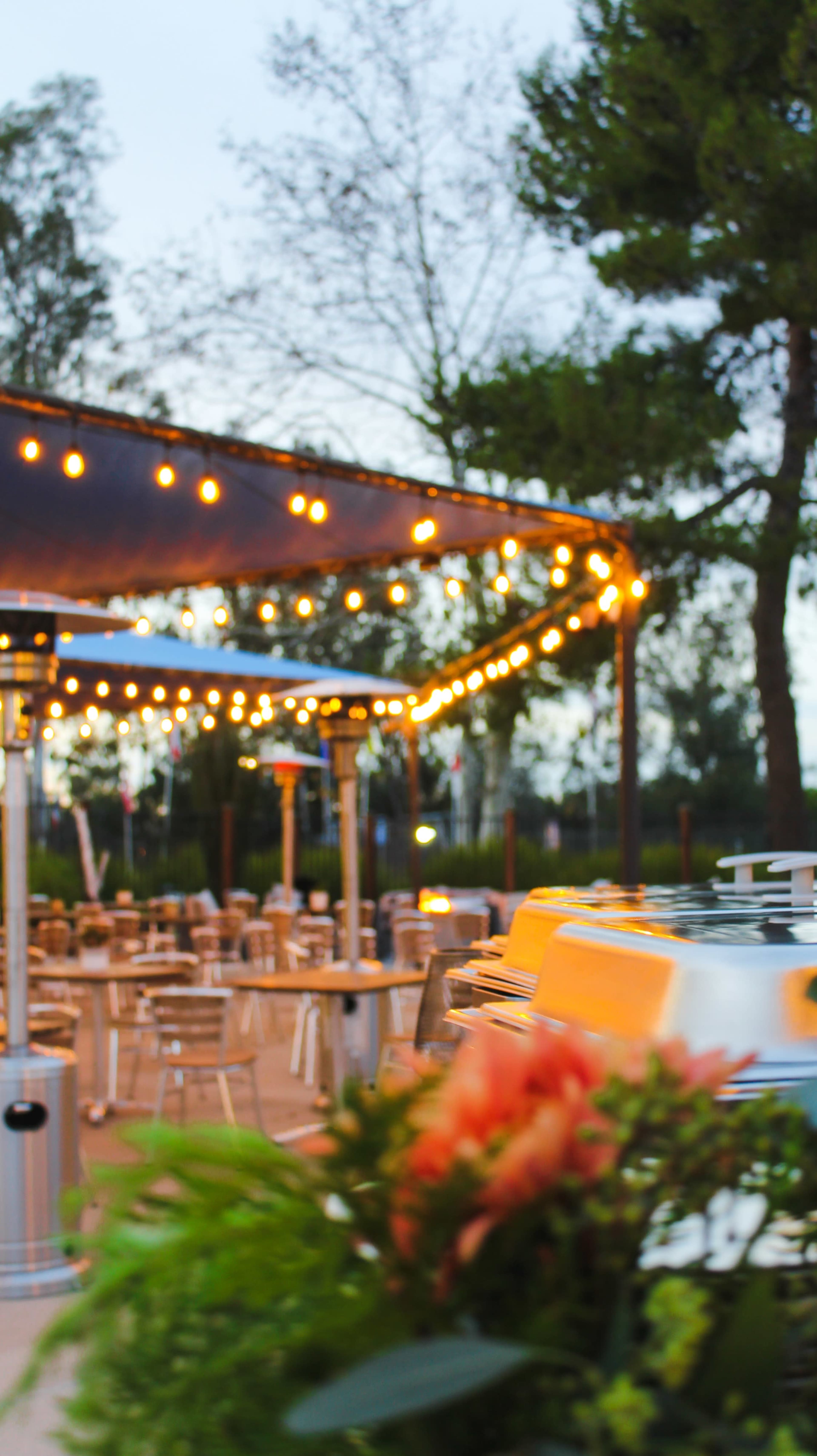 An outdoor dining area illuminated by string lights, with tables and chairs arranged under a canopy.