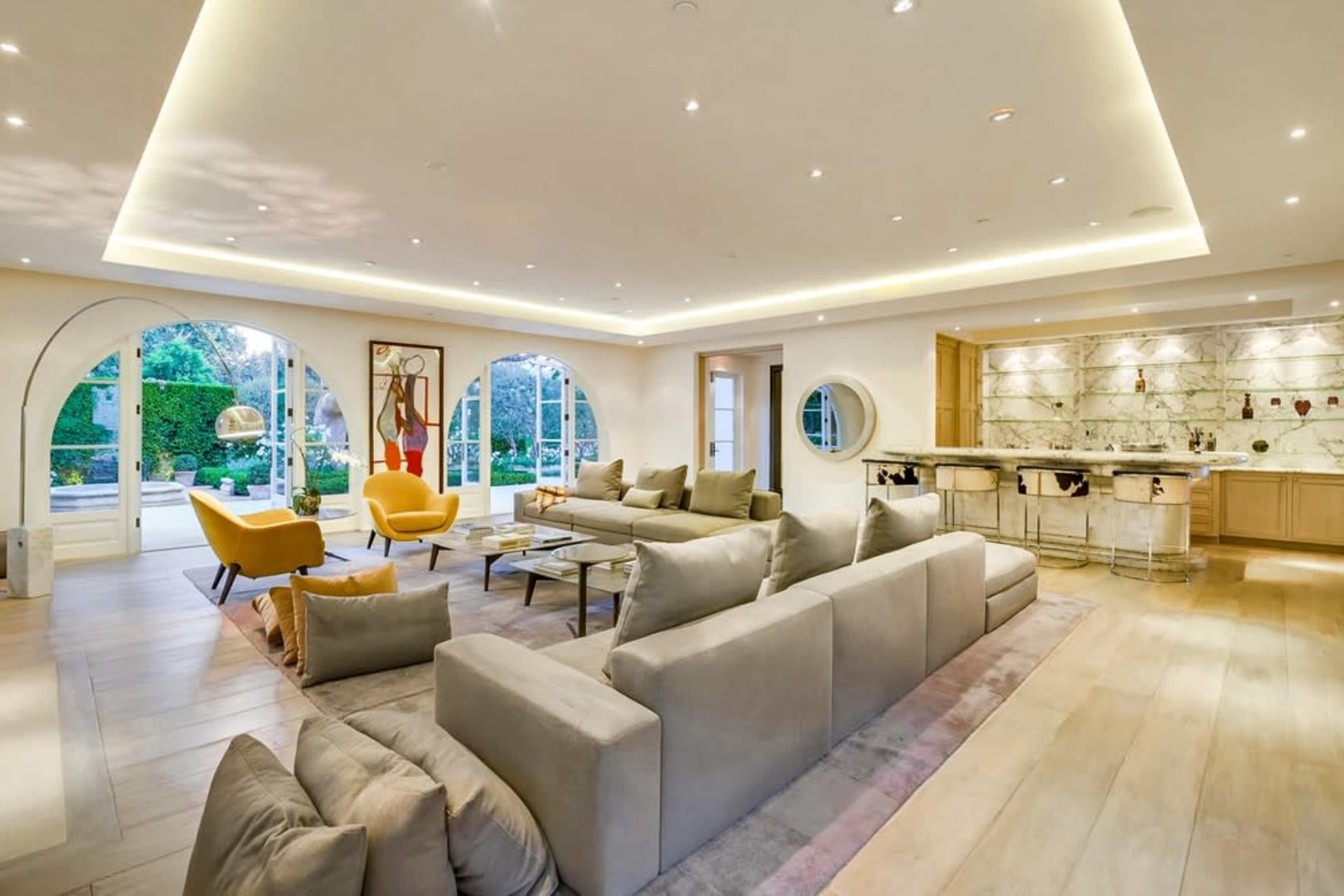 A modern living room with a large sectional sofa, yellow armchairs, and a sleek kitchen in the background, illuminated by recessed lighting and large windows that allow natural light.