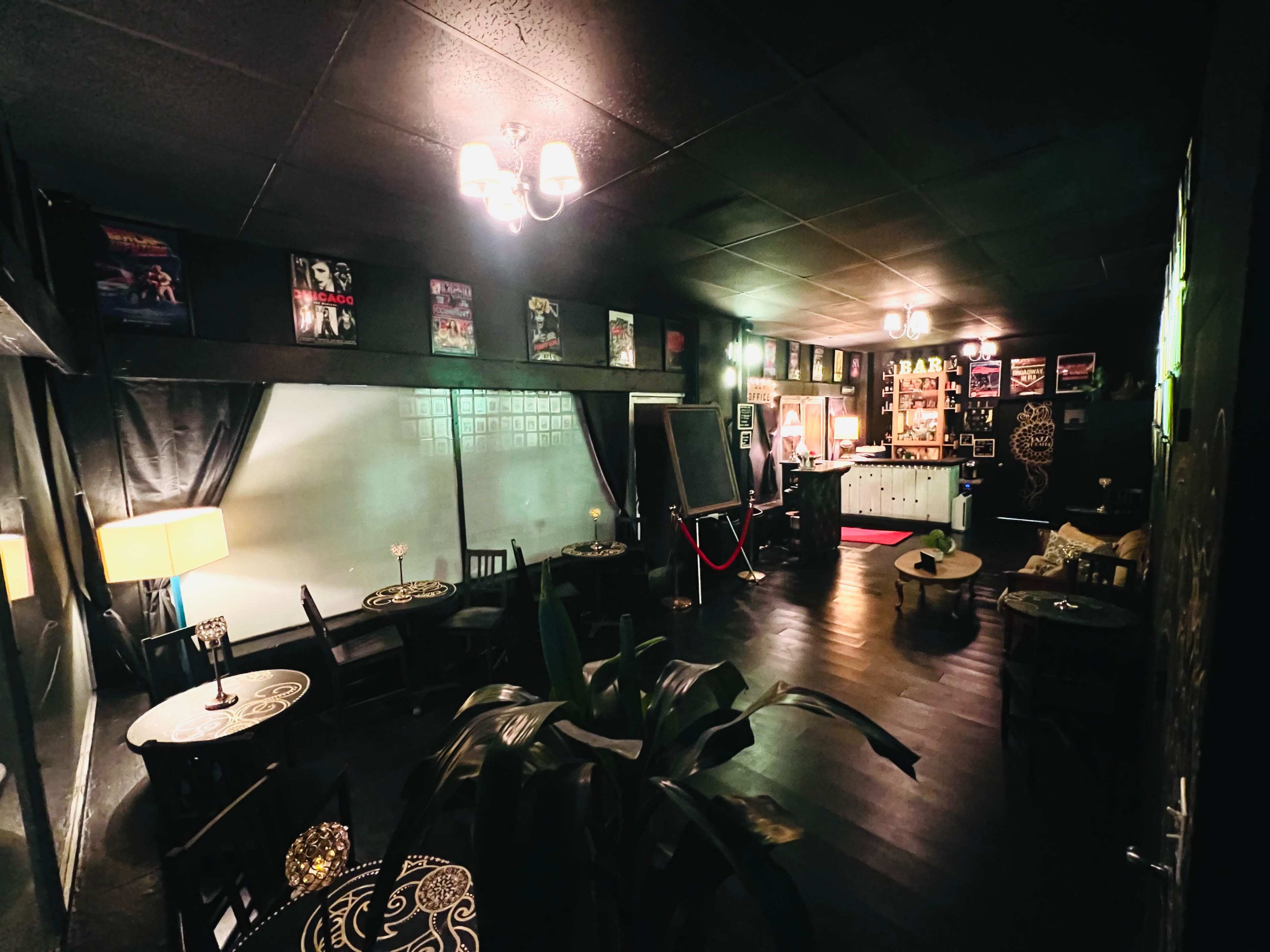 The image shows a dimly lit bar with black walls, tables arranged for seating, and a well-stocked bar counter in the background.