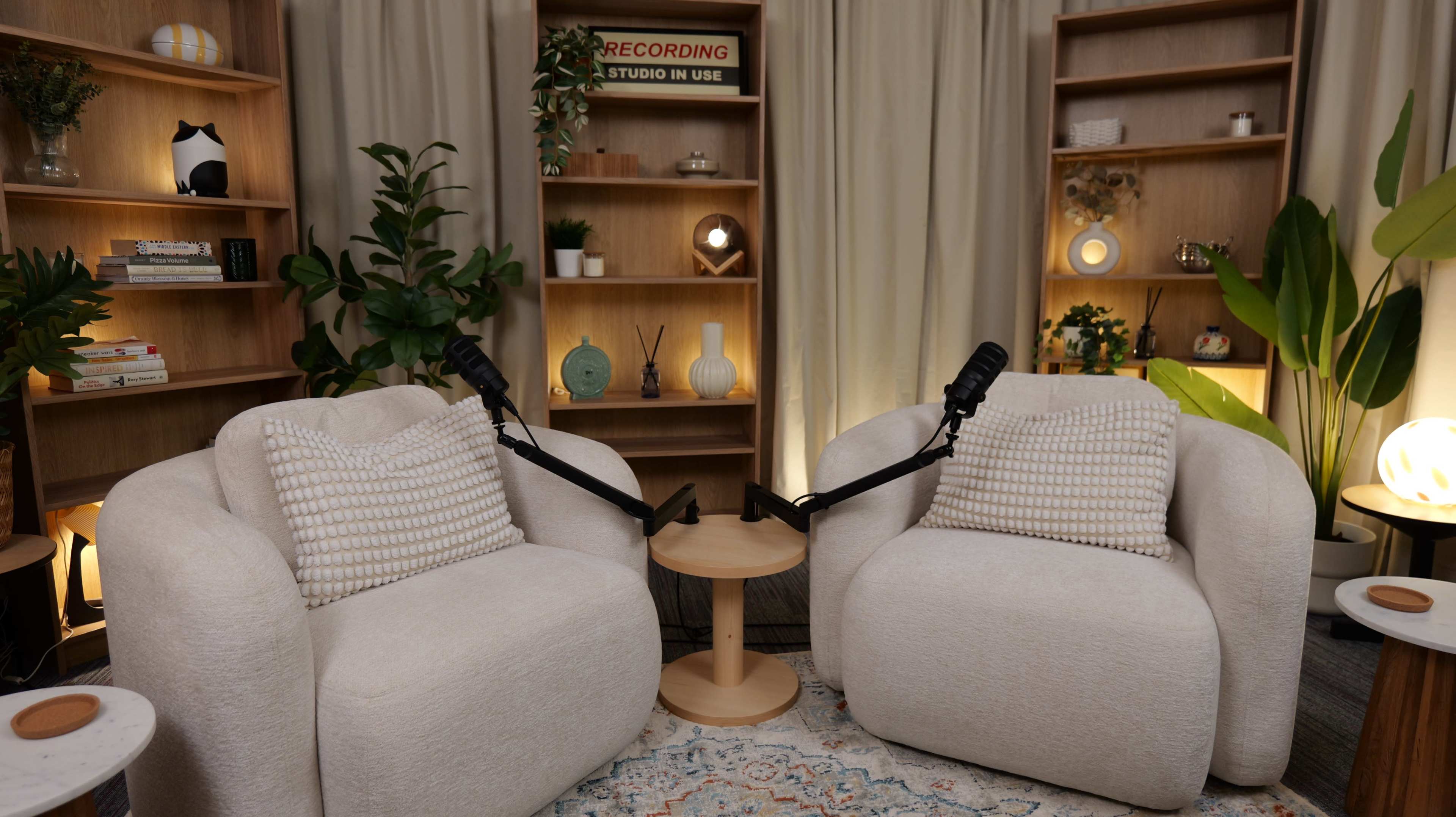 The image shows a cozy recording studio setup with two plush armchairs facing each other, a small table between them, and a backdrop of wooden shelves adorned with decorative items and plants.
