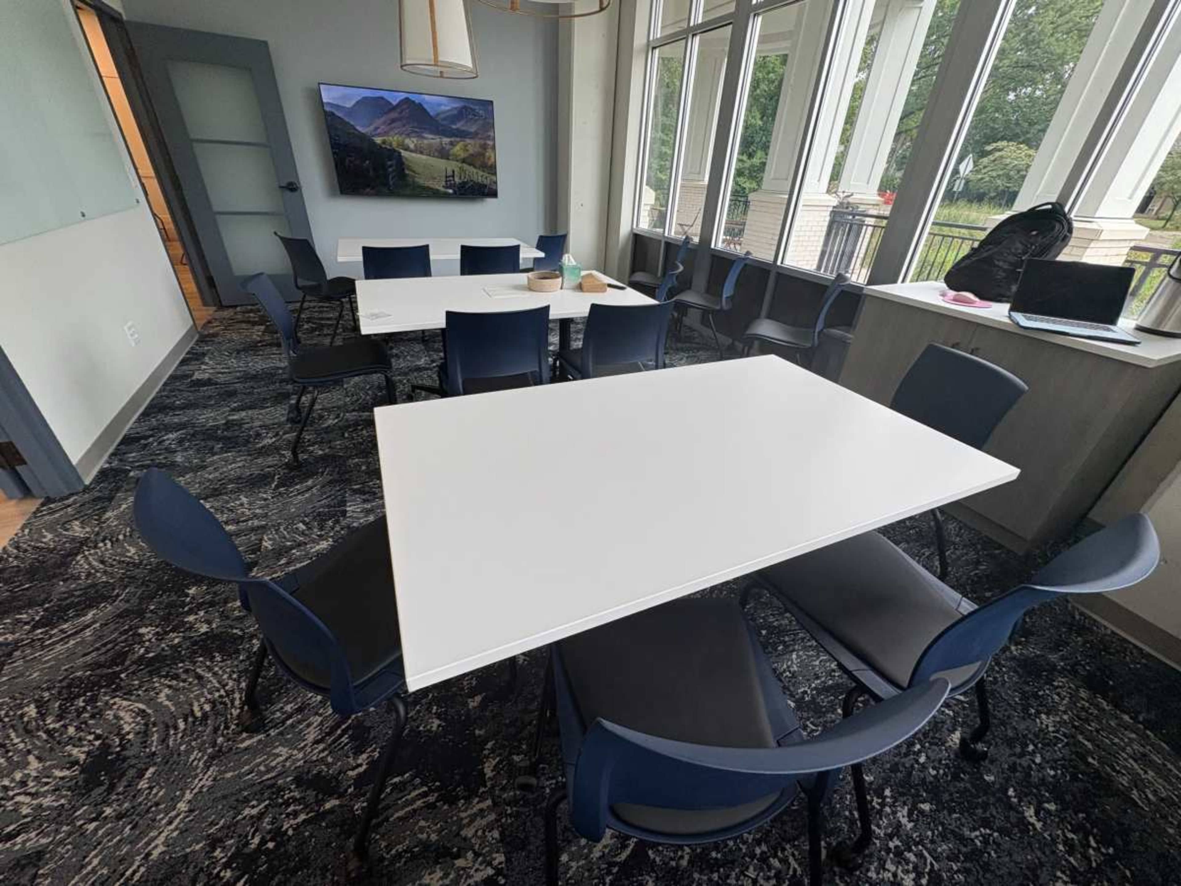 A modern conference room features a large white table surrounded by several blue chairs, with a view of outdoor greenery through large windows.