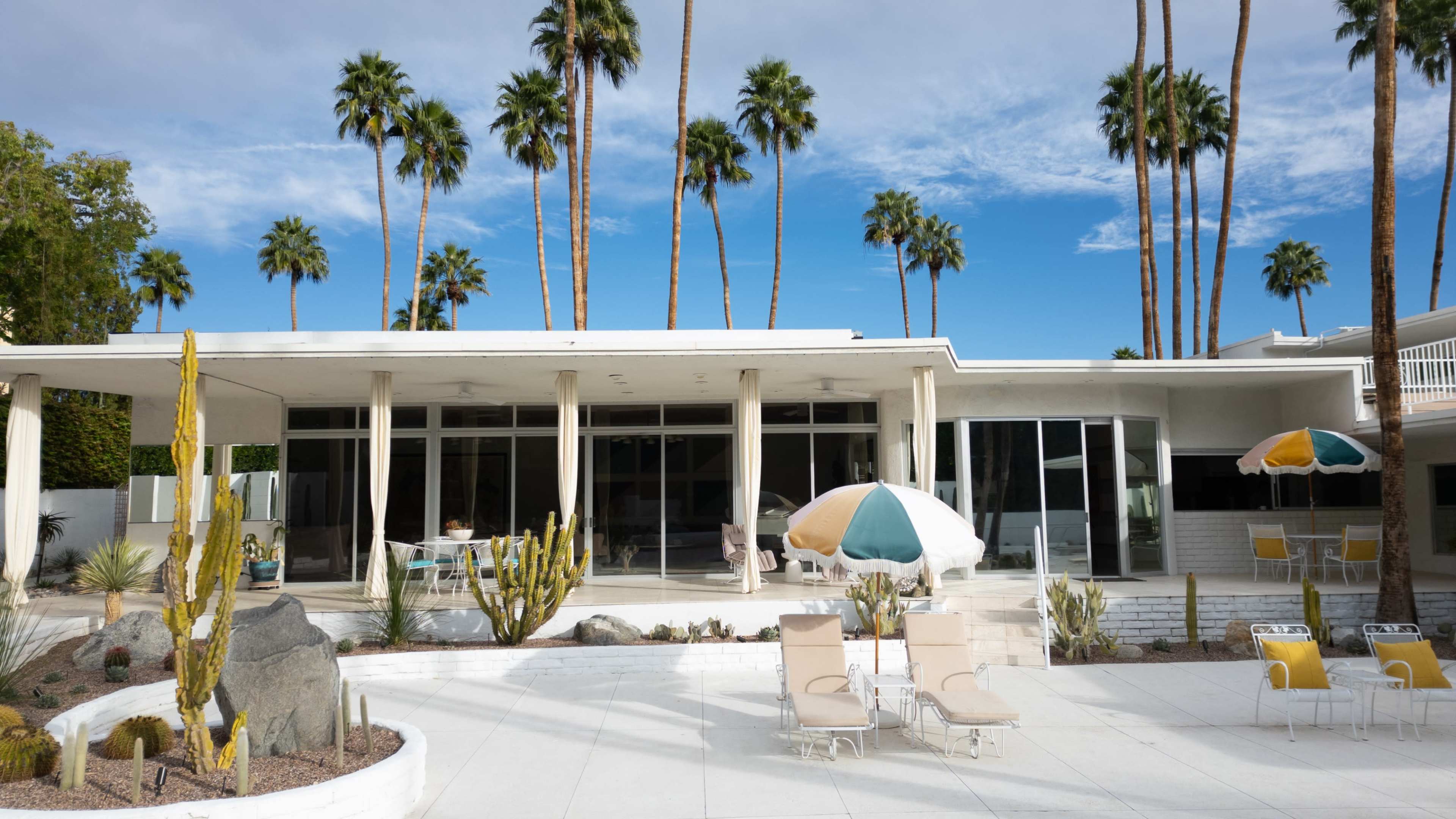 The image features a modern, single-story building with large glass windows, surrounded by palm trees and a landscaped patio area with lounge chairs and umbrellas.