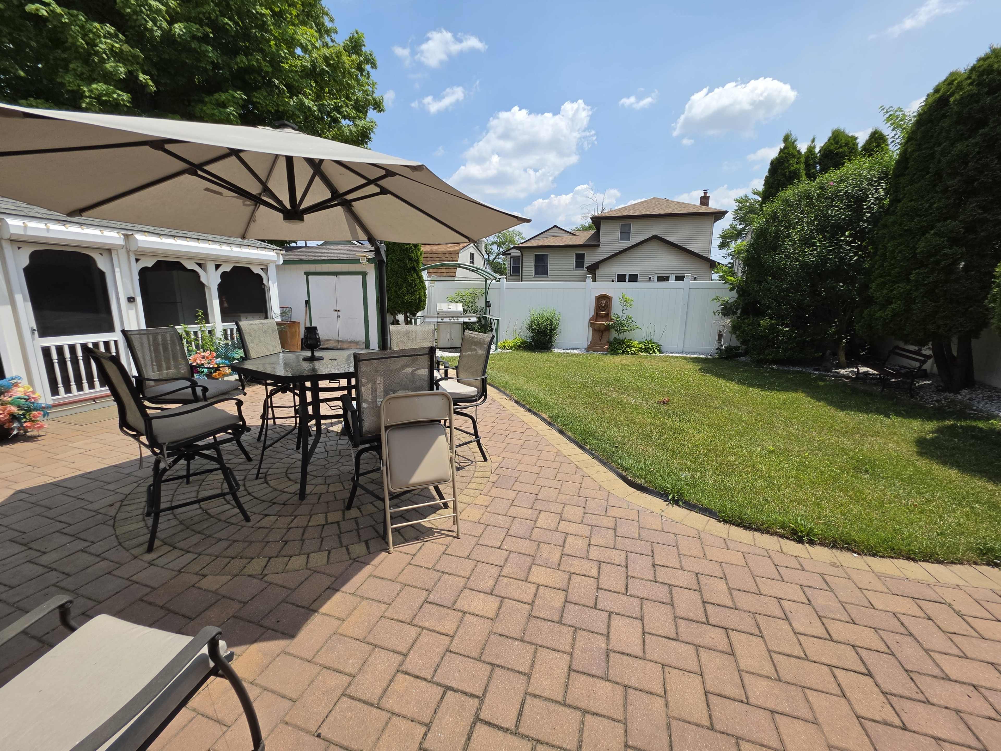 The image shows a backyard patio with a round table and chairs beneath a large umbrella, surrounded by green grass and a fence in the background.
