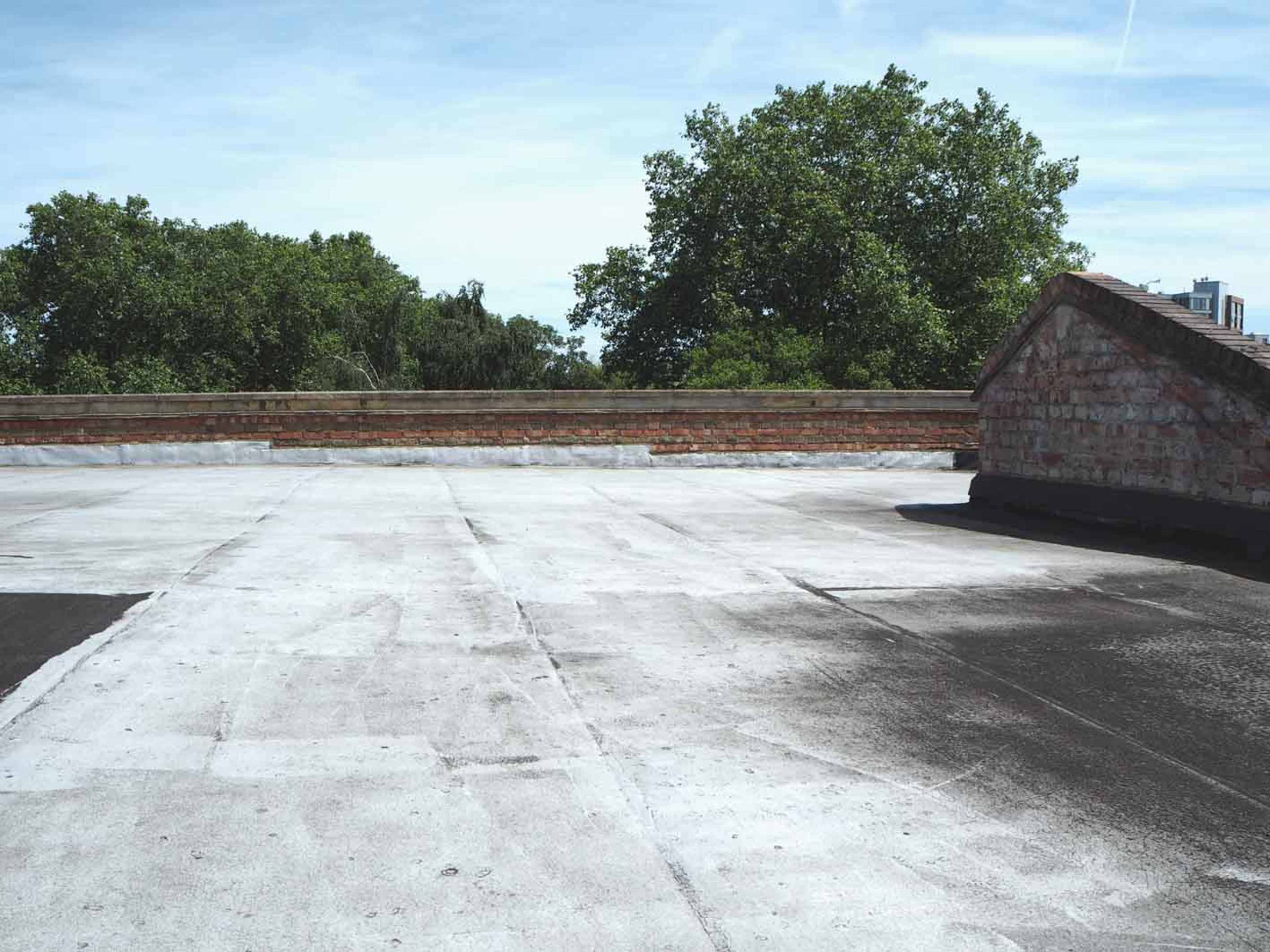 A flat rooftop surface with a light-colored finish, surrounded by greenery and a brick wall.