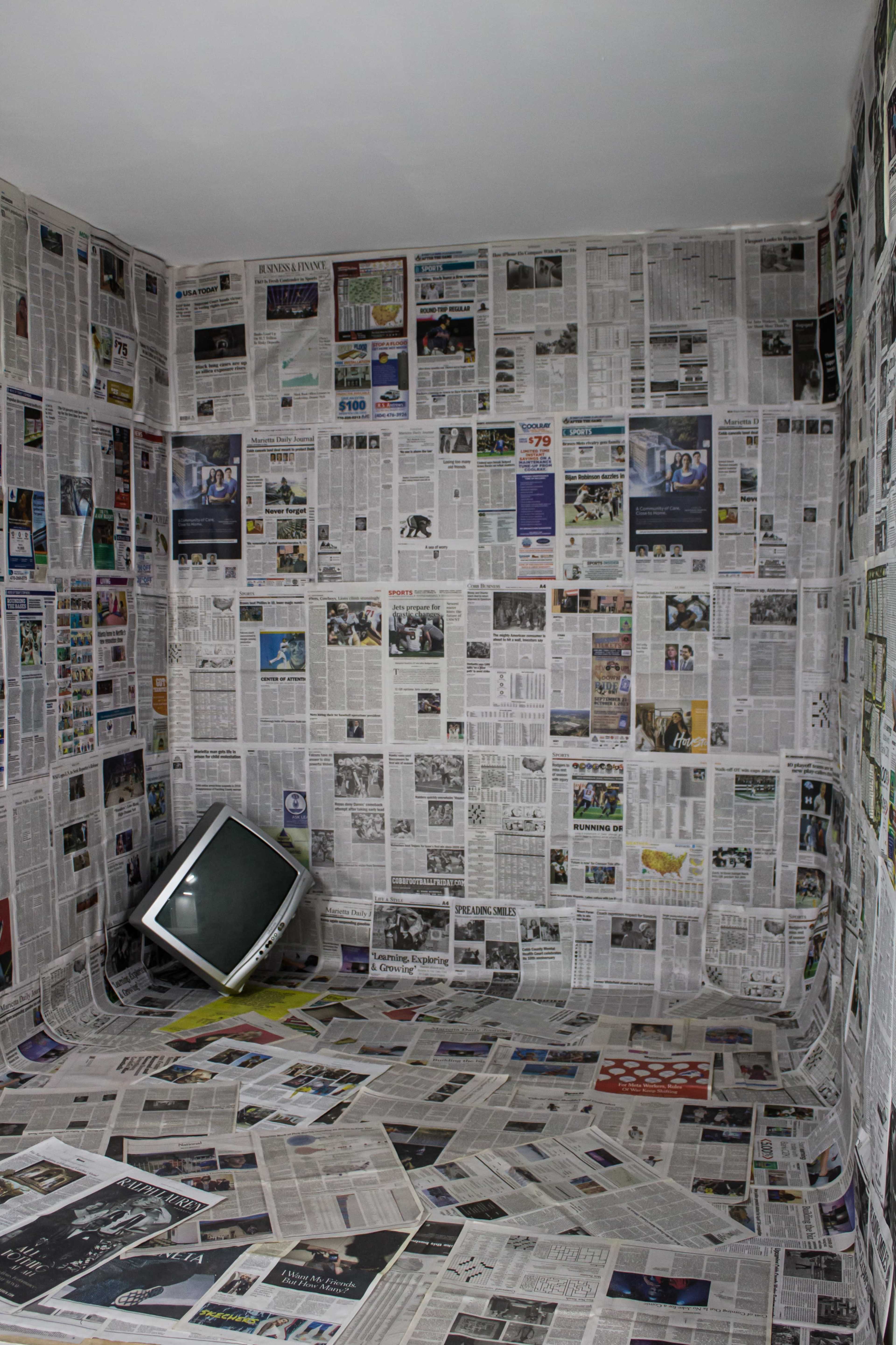 A small room is entirely covered with newspapers, including the walls, floor, and an old television placed in one corner.