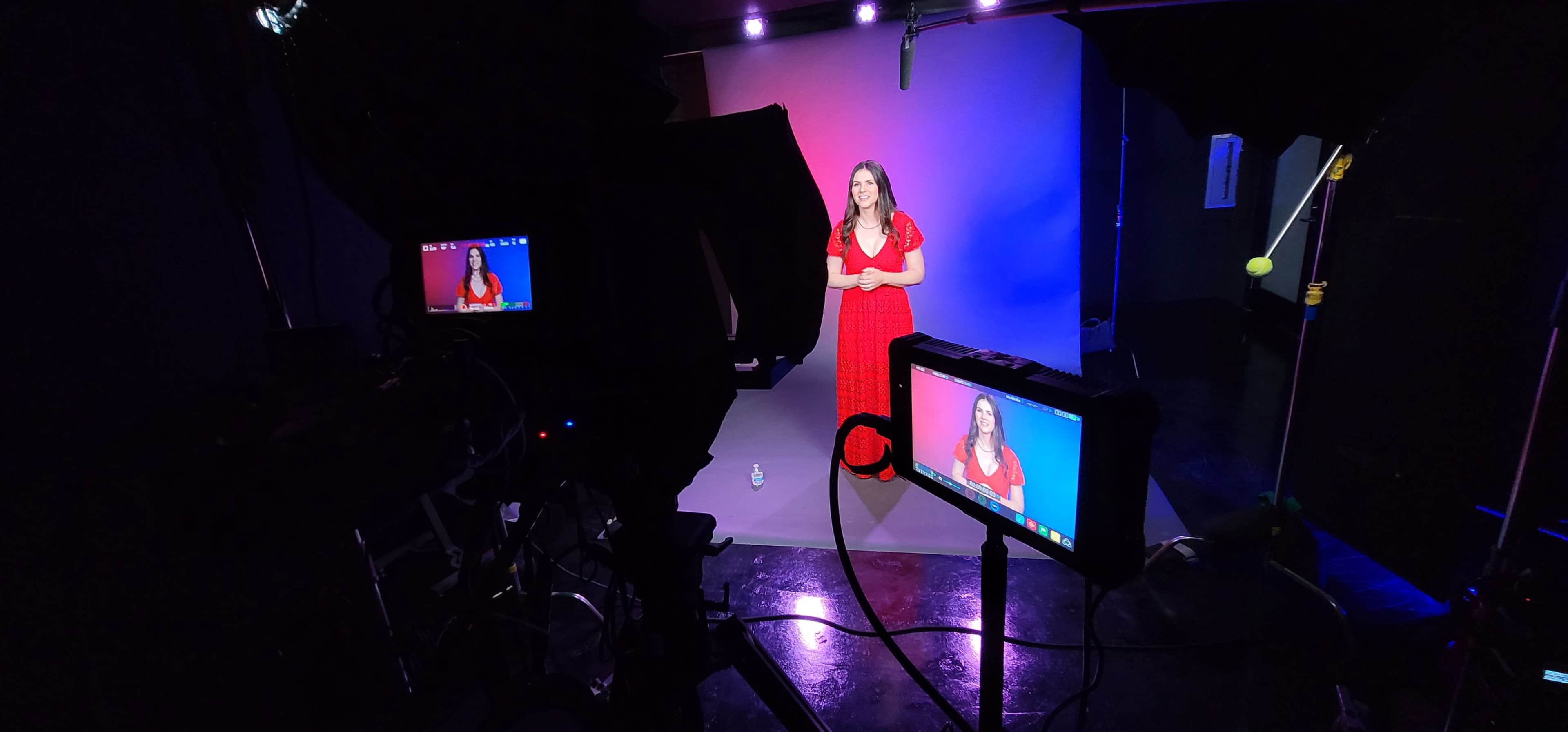 A woman in a red dress stands in front of a blue and purple backdrop while being filmed in a studio setting.