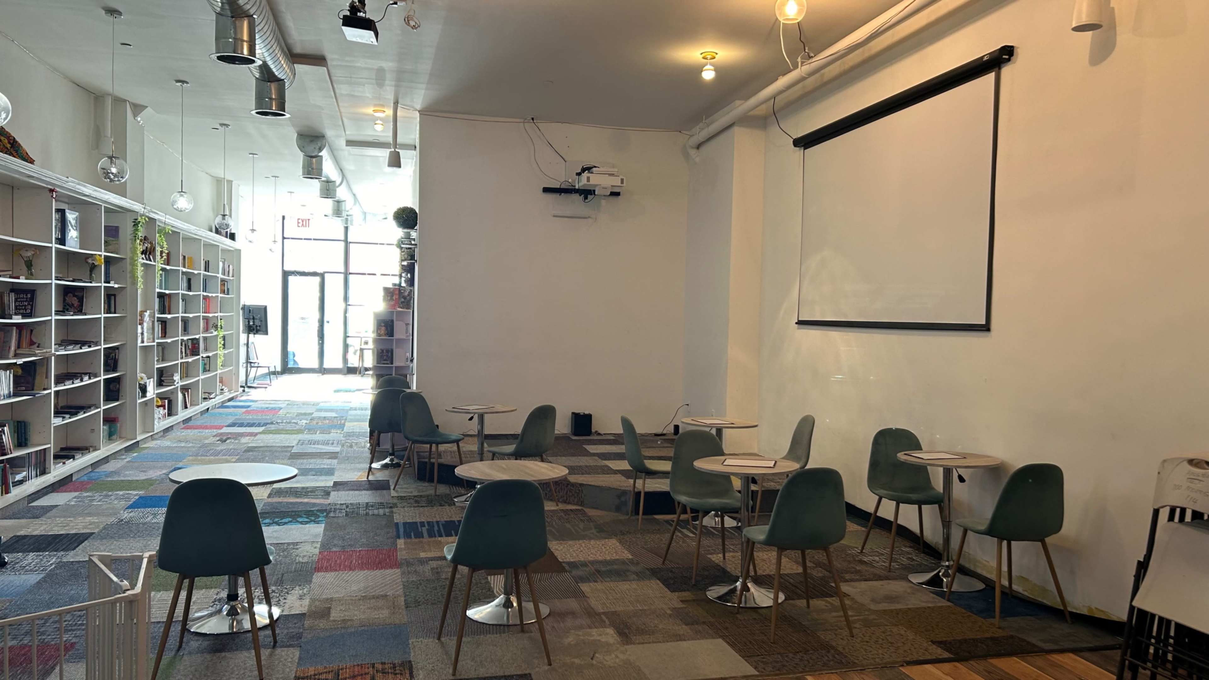A spacious cafe area featuring several small round tables and chairs, with a large blank screen on one wall and shelves filled with books.