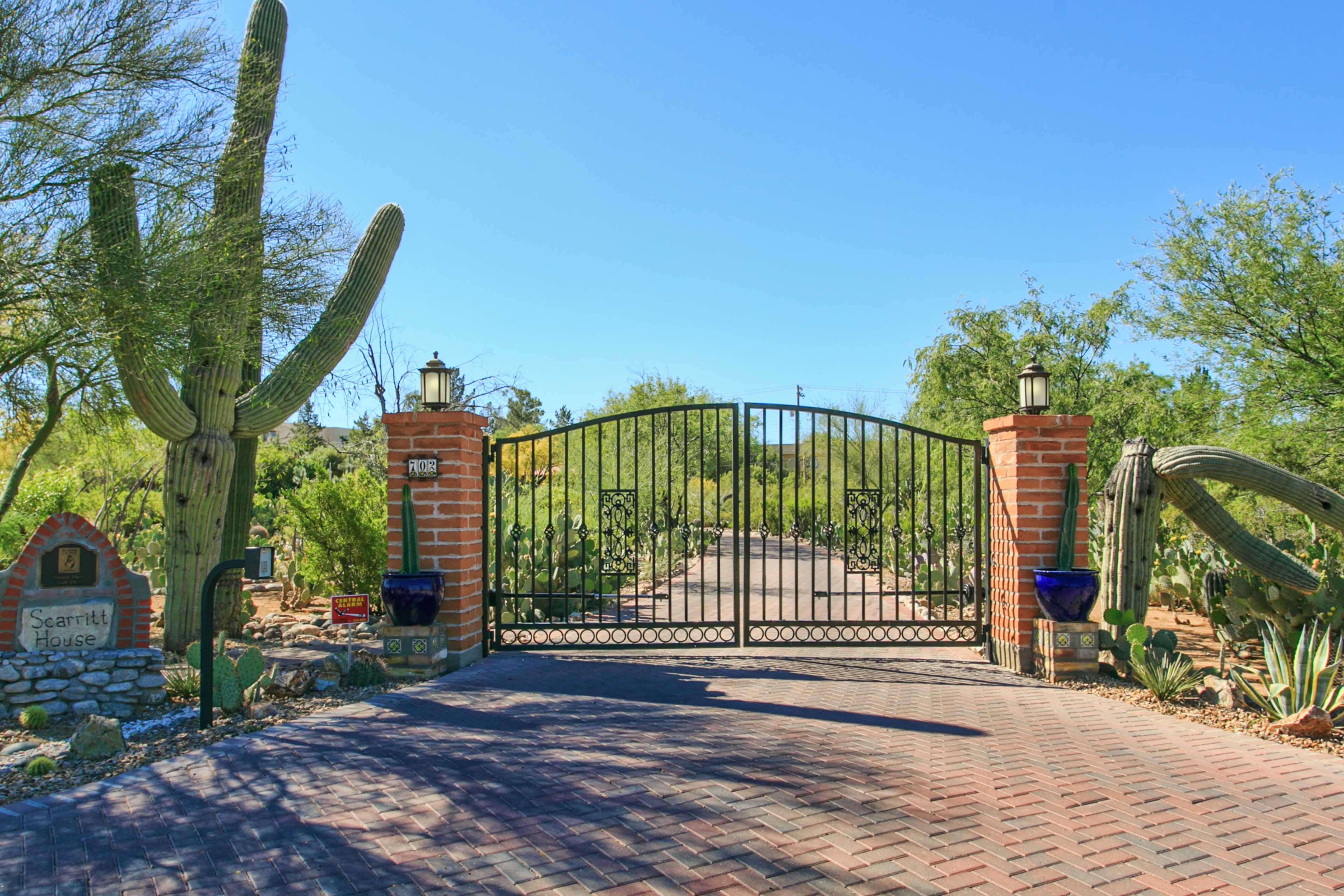 A wrought iron gate framed by brick columns opens into a landscaped pathway lined with desert plants and cacti.
