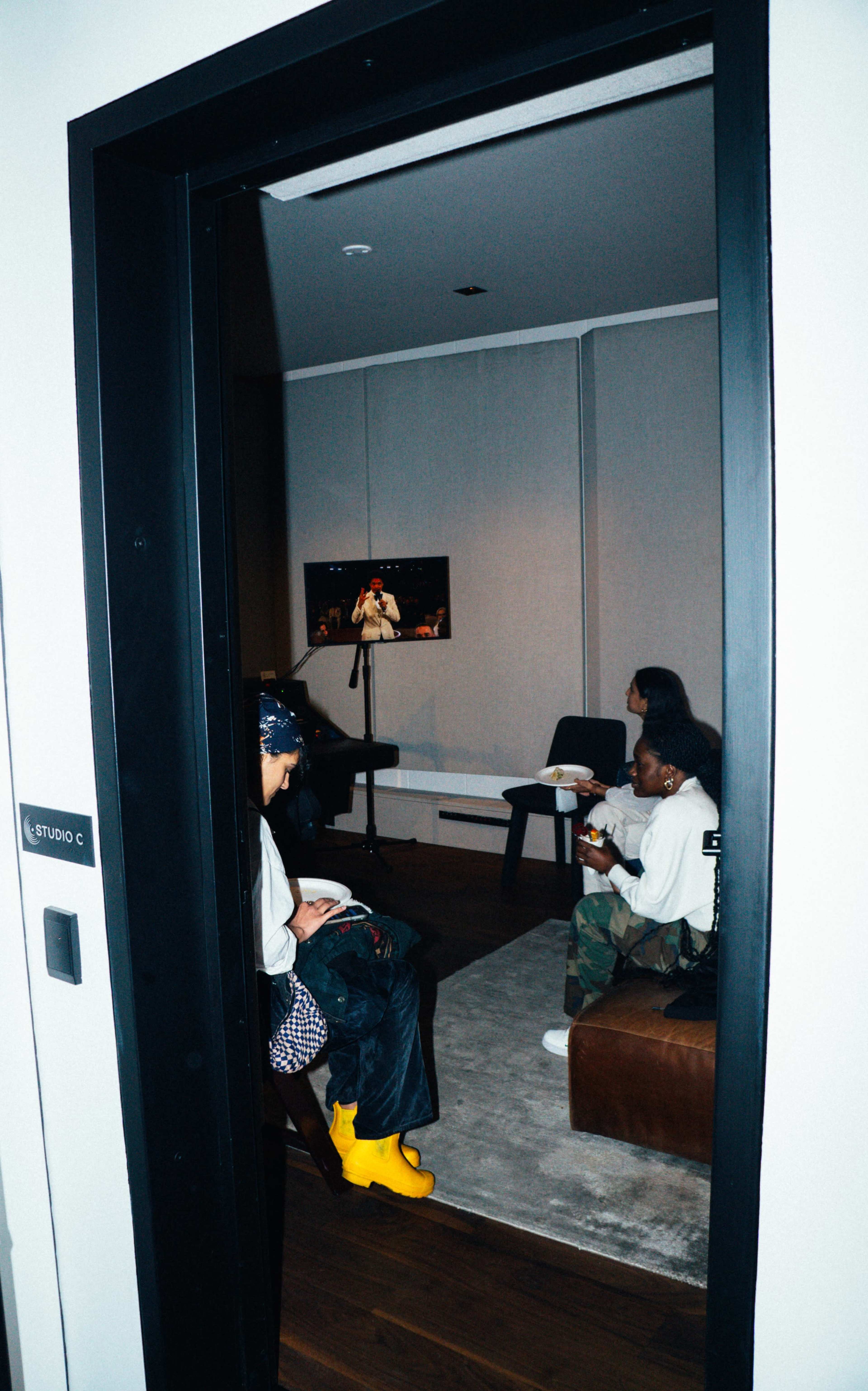 Three people sit in a room, watching a performance on a screen while seated on a couch and chairs.
