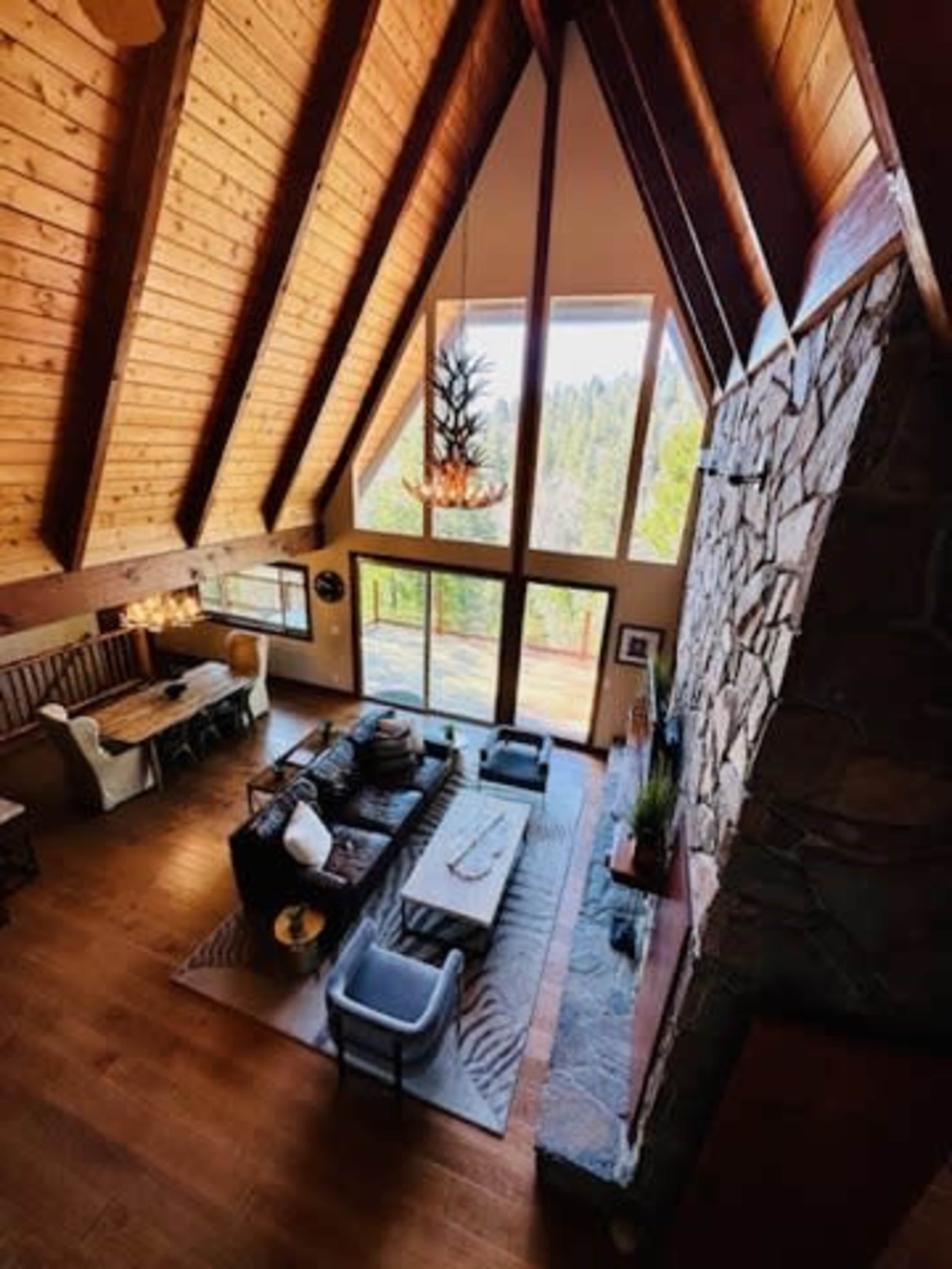A spacious, open-concept living area features a high, wooden A-frame ceiling, large windows, and a stone fireplace, complemented by contemporary furniture.