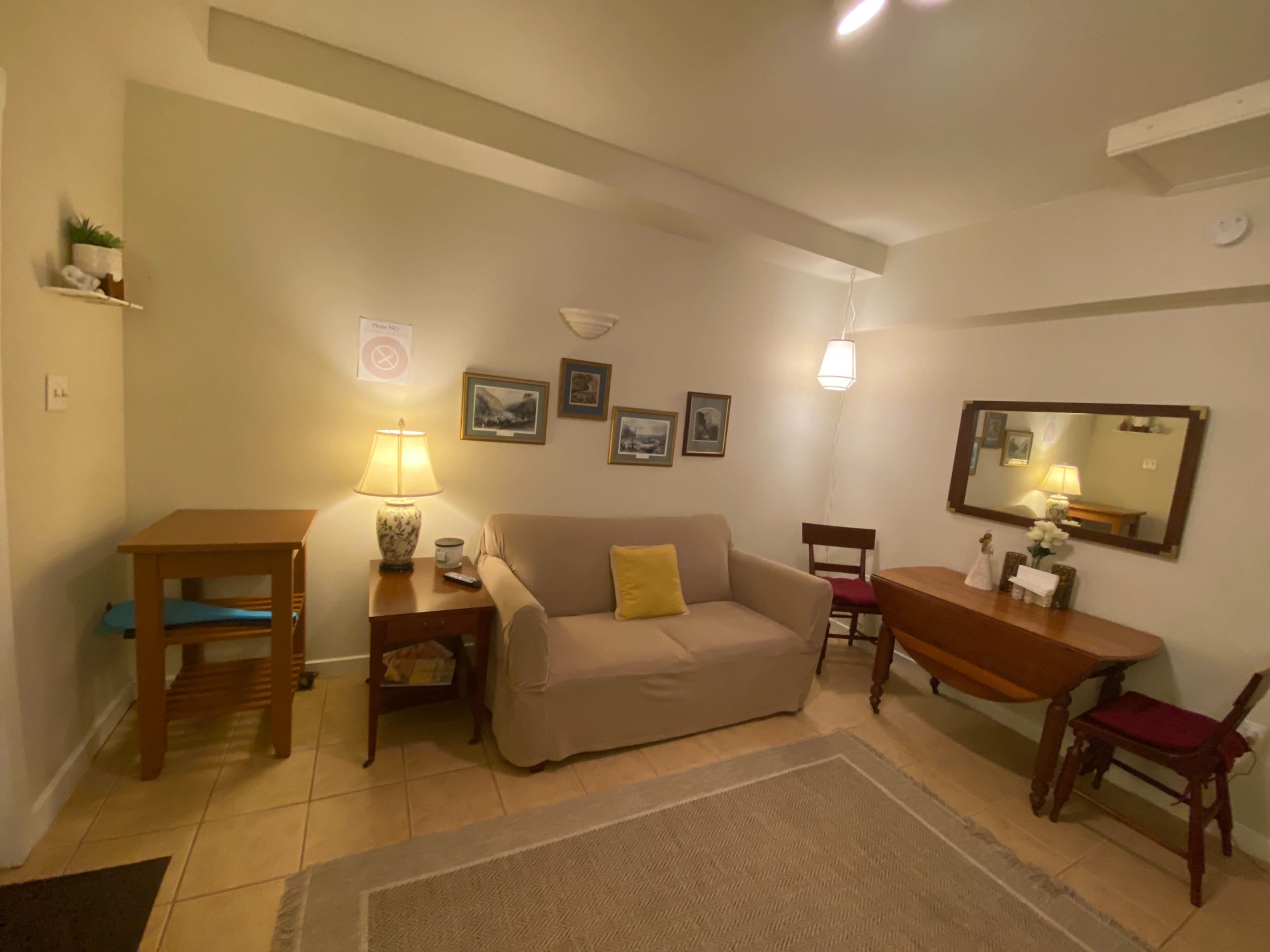 The image shows a small living area with a sofa, a wooden table, a lamp, and decorative wall art.