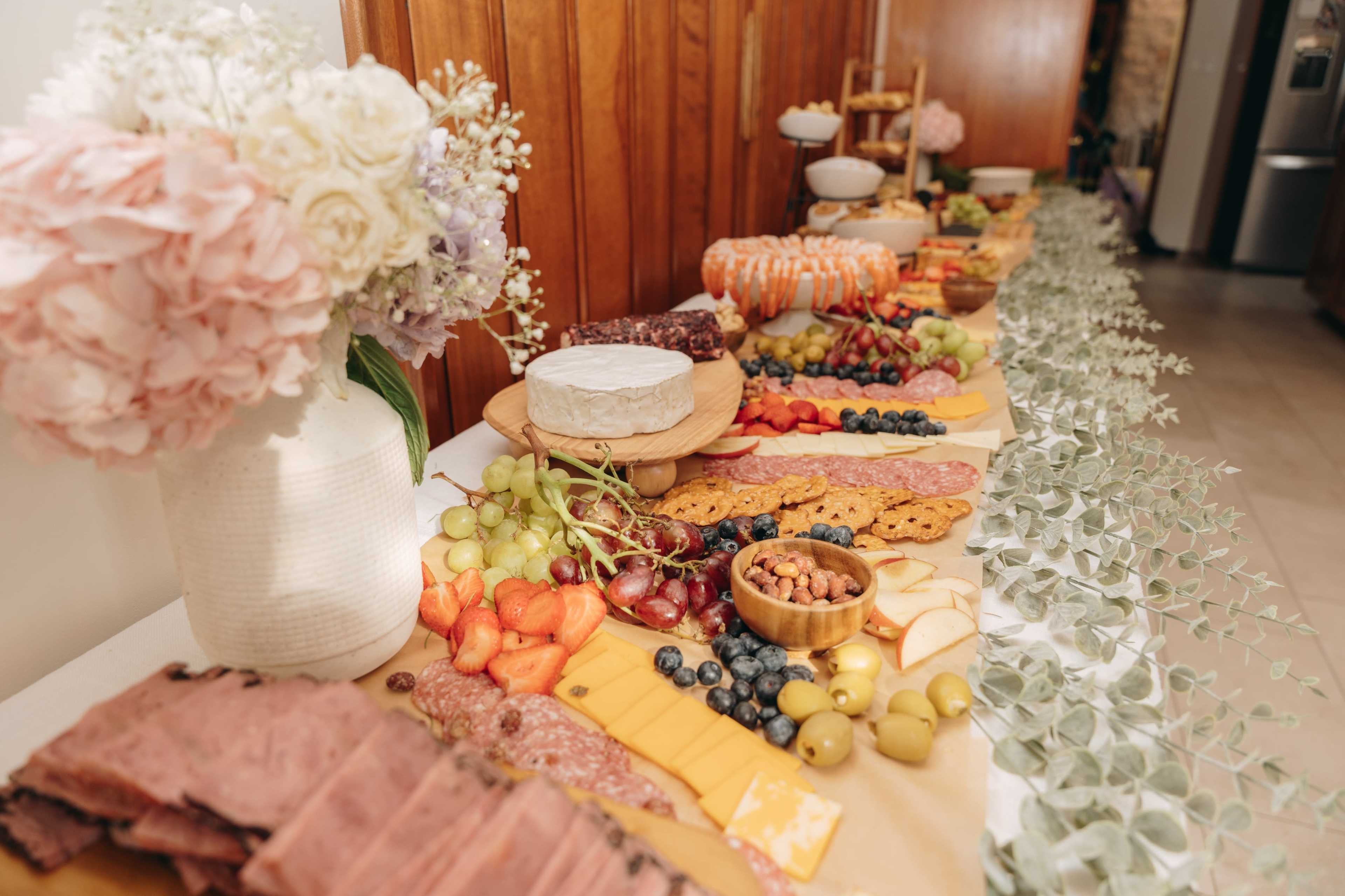 A long table is set with an assortment of meats, cheeses, fruits, crackers, and flowers arranged along the center.
