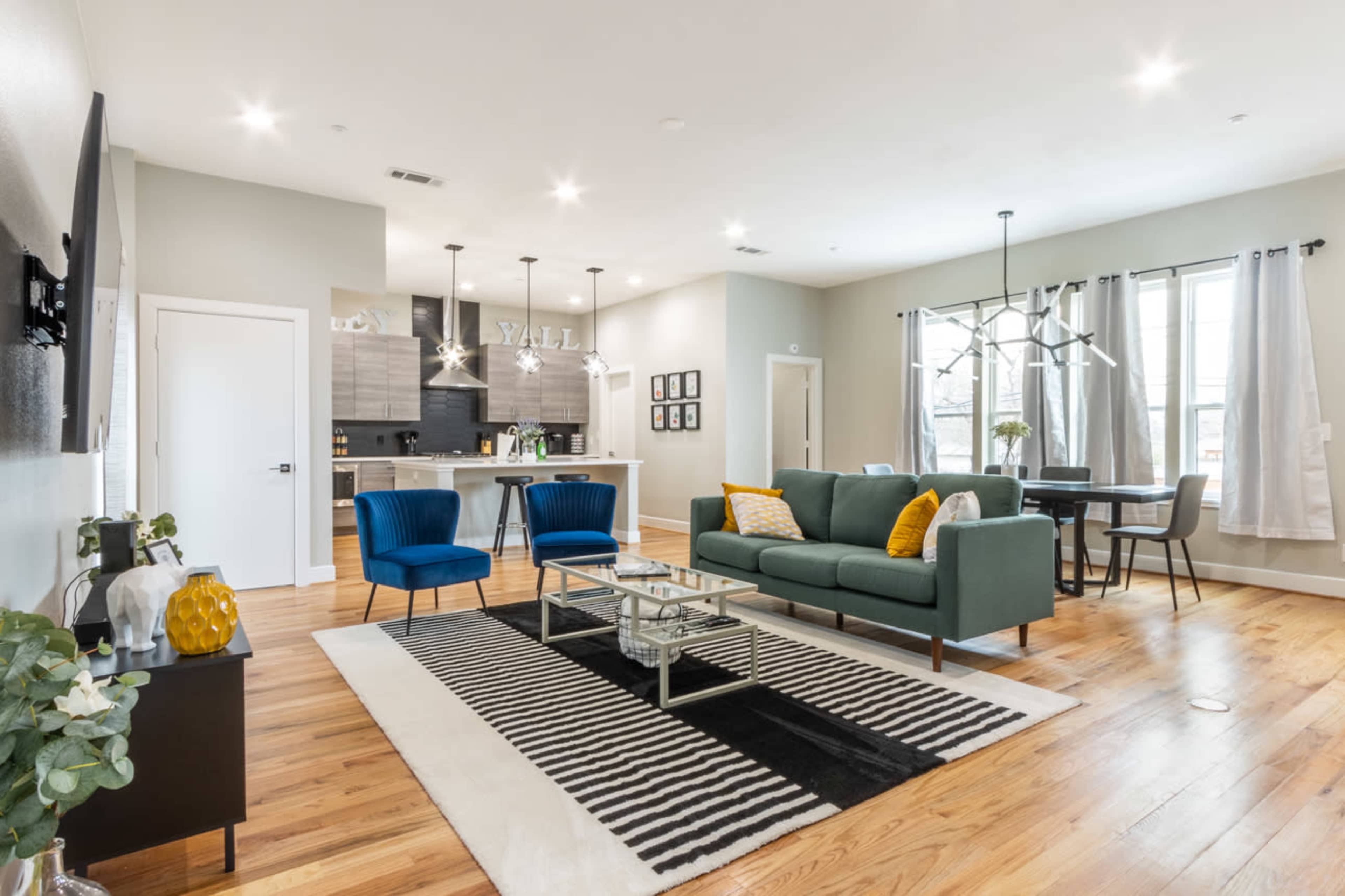 A modern living room with a green sofa, two blue chairs, a coffee table, and wooden flooring, along with an adjacent kitchen area visible in the background.