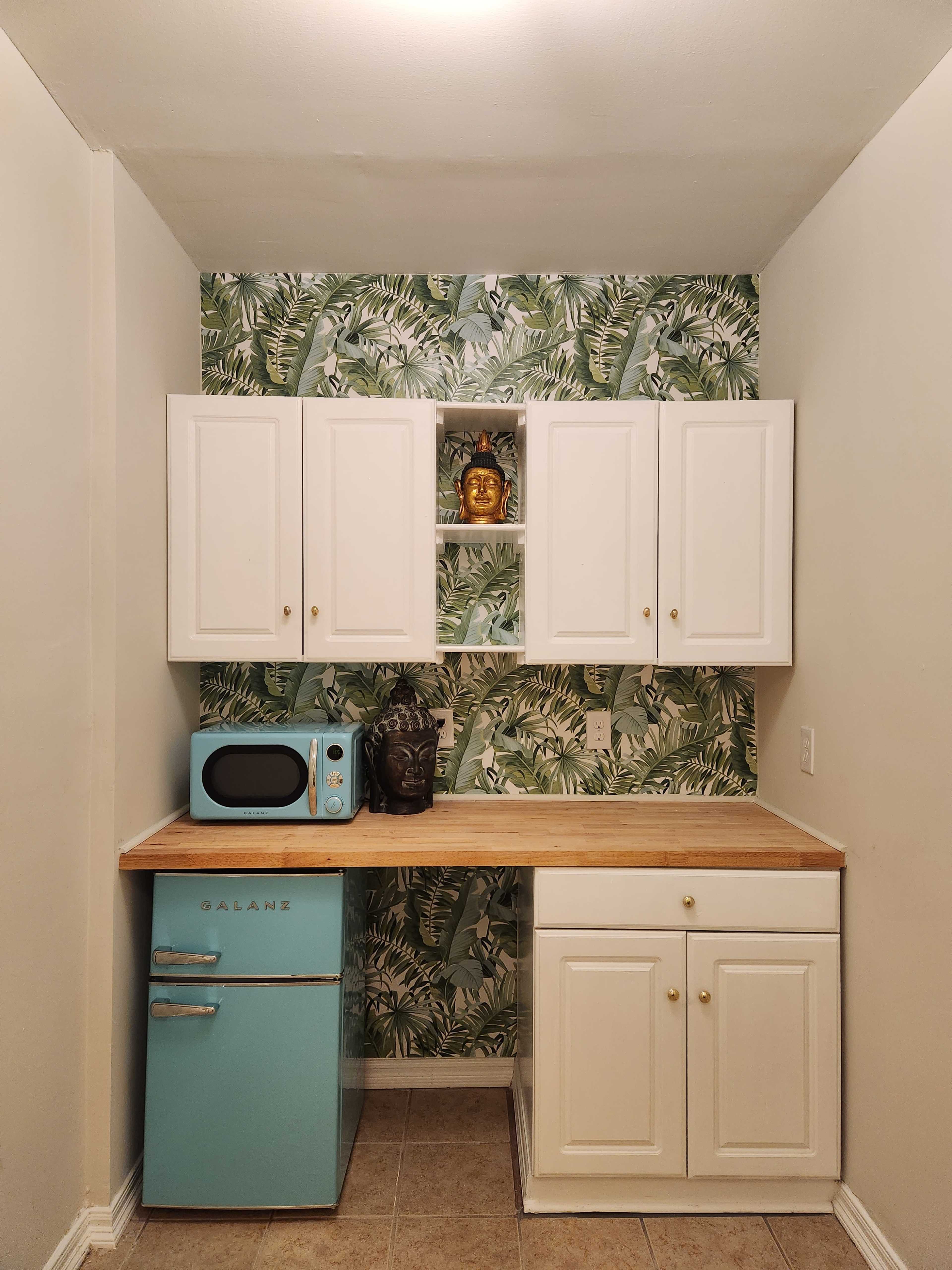 The image shows a small kitchen area featuring a light blue fridge, white cabinets, a microwave, and a patterned green wallpaper with palm leaves.
